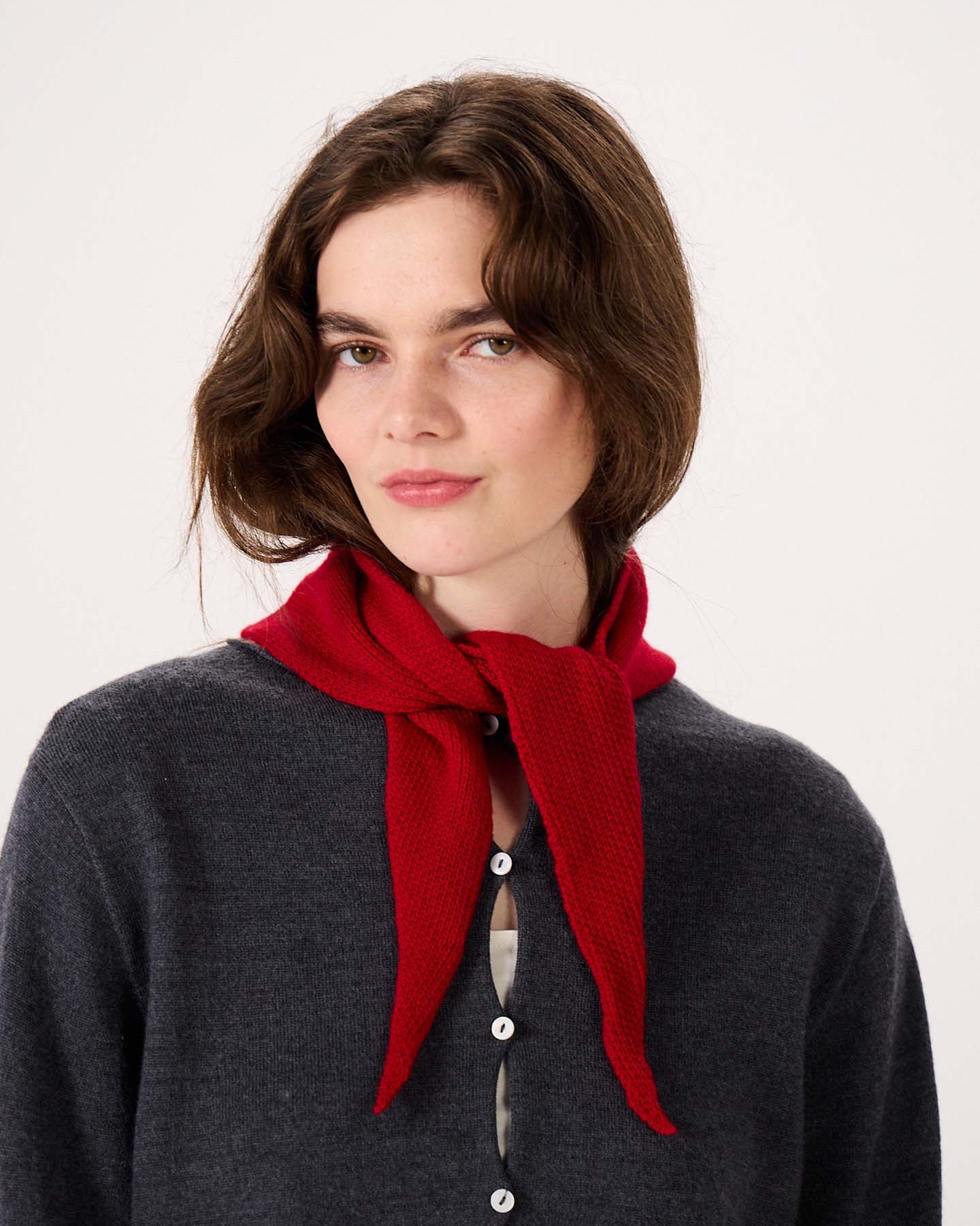 A woman with wavy brown hair is wearing a dark gray buttoned sweater and the Daisy Scarf – Tamarillo, tied around her neck, standing against a plain light background. The scarf measures 97cm long and 22cm wide, offering stylish comfort with practical product dimensions.