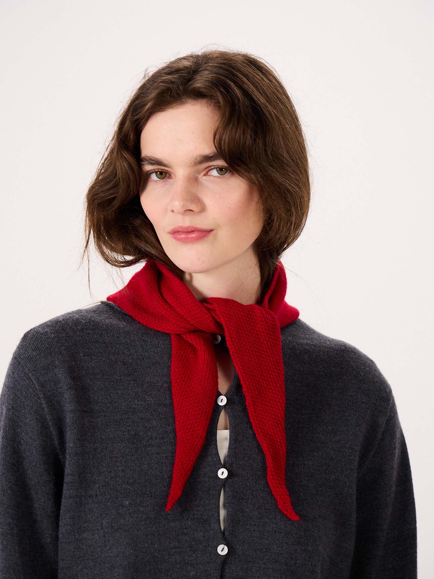 A woman with wavy brown hair is wearing a dark gray buttoned sweater and the Daisy Scarf – Tamarillo, tied around her neck, standing against a plain light background. The scarf measures 97cm long and 22cm wide, offering stylish comfort with practical product dimensions.