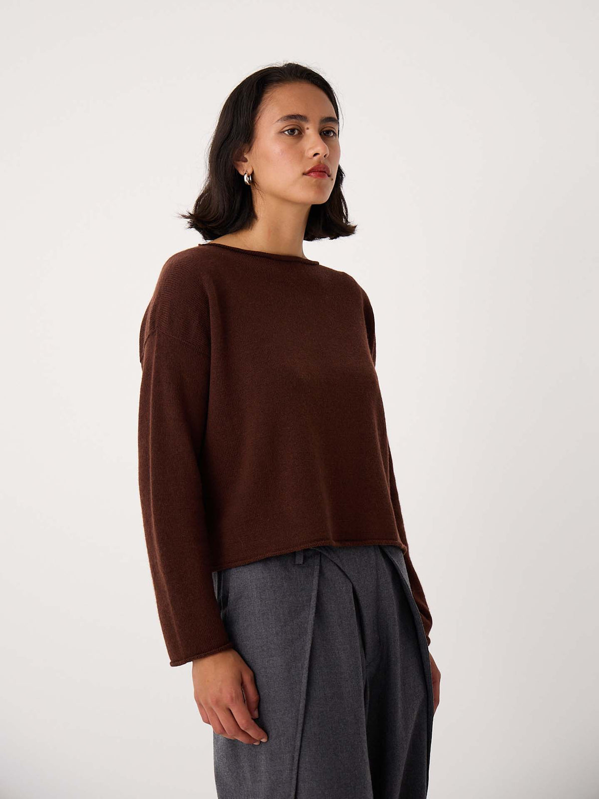 A person with shoulder-length dark hair wears the Doll's Jumper – Chestnut, a relaxed fit brown long-sleeve sweater, with gray pleated pants, standing against a plain light background.