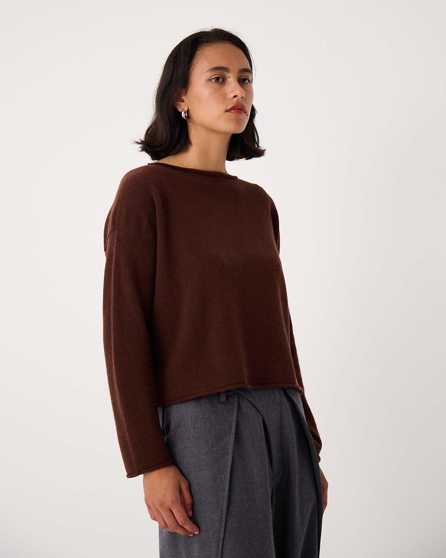 A person with shoulder-length dark hair wears the Doll's Jumper – Chestnut, a relaxed fit brown long-sleeve sweater, with gray pleated pants, standing against a plain light background.