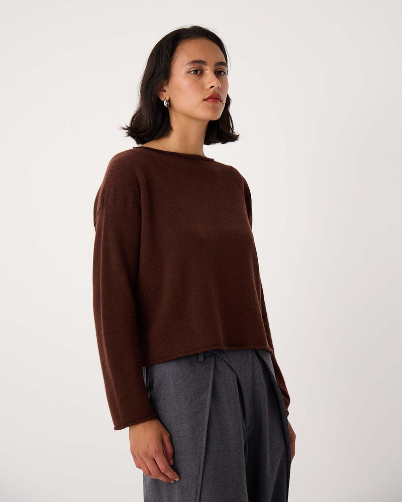 A person with shoulder-length dark hair wears the Doll's Jumper – Chestnut, a relaxed fit brown long-sleeve sweater, with gray pleated pants, standing against a plain light background.