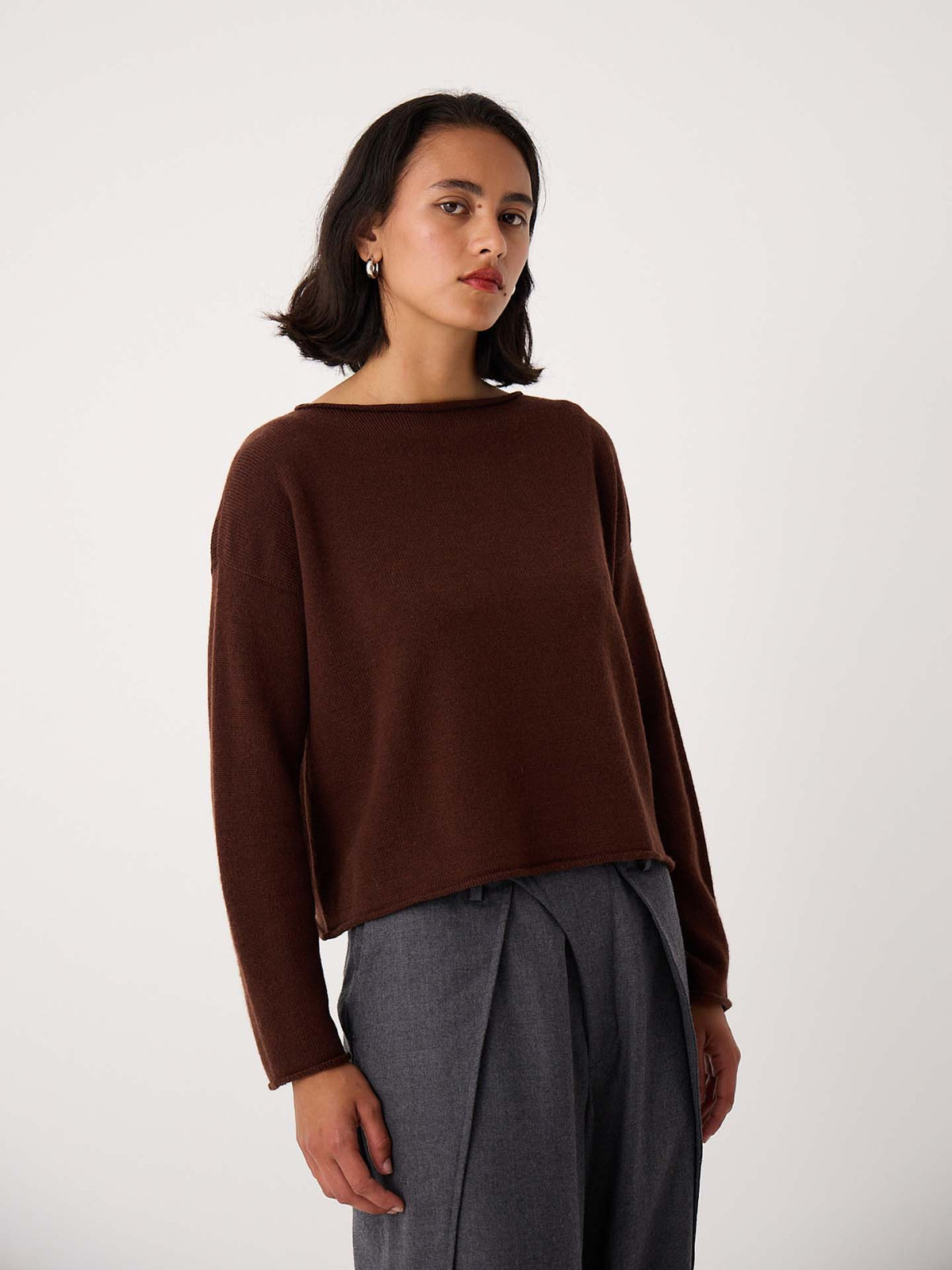 A person with shoulder-length dark hair wears the Doll's Jumper – Chestnut, a brown relaxed fit long-sleeve top, and gray pleated trousers, standing against a plain light background.