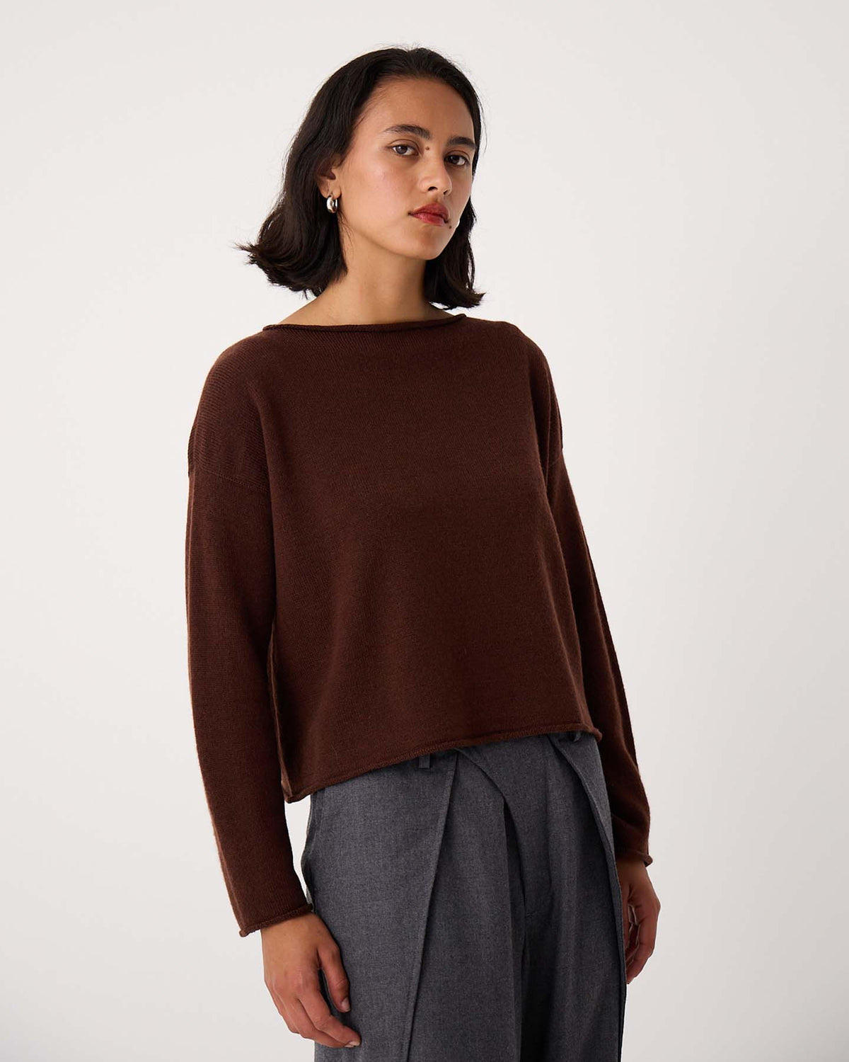 A person with shoulder-length dark hair wears the Doll's Jumper – Chestnut, a brown relaxed fit long-sleeve top, and gray pleated trousers, standing against a plain light background.