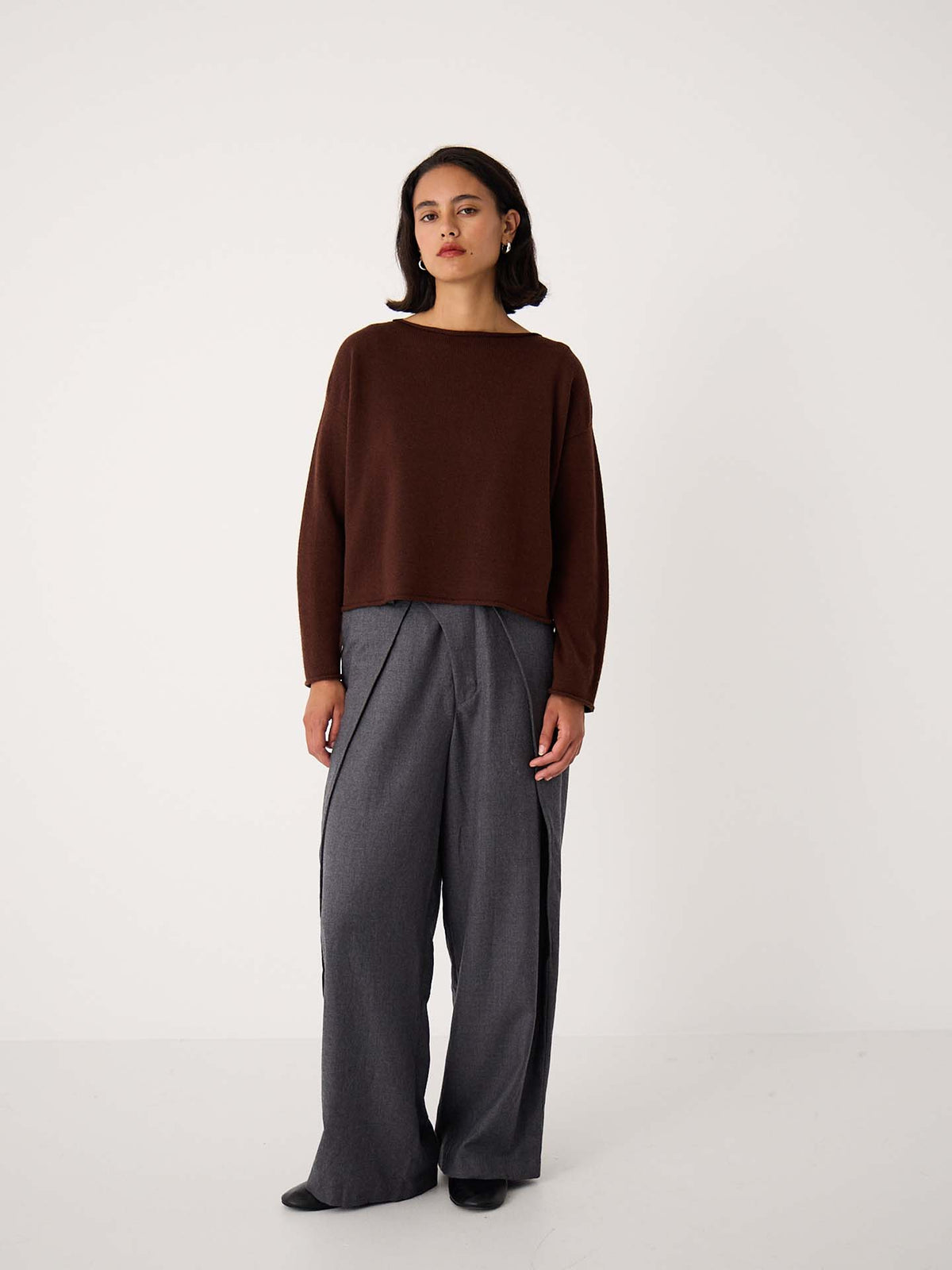 A woman stands against a plain white background wearing the Doll's Jumper – Chestnut, a relaxed fit cropped brown sweater, paired with wide-leg gray trousers and black shoes—an effortless look ideal for petite clothing lovers.
