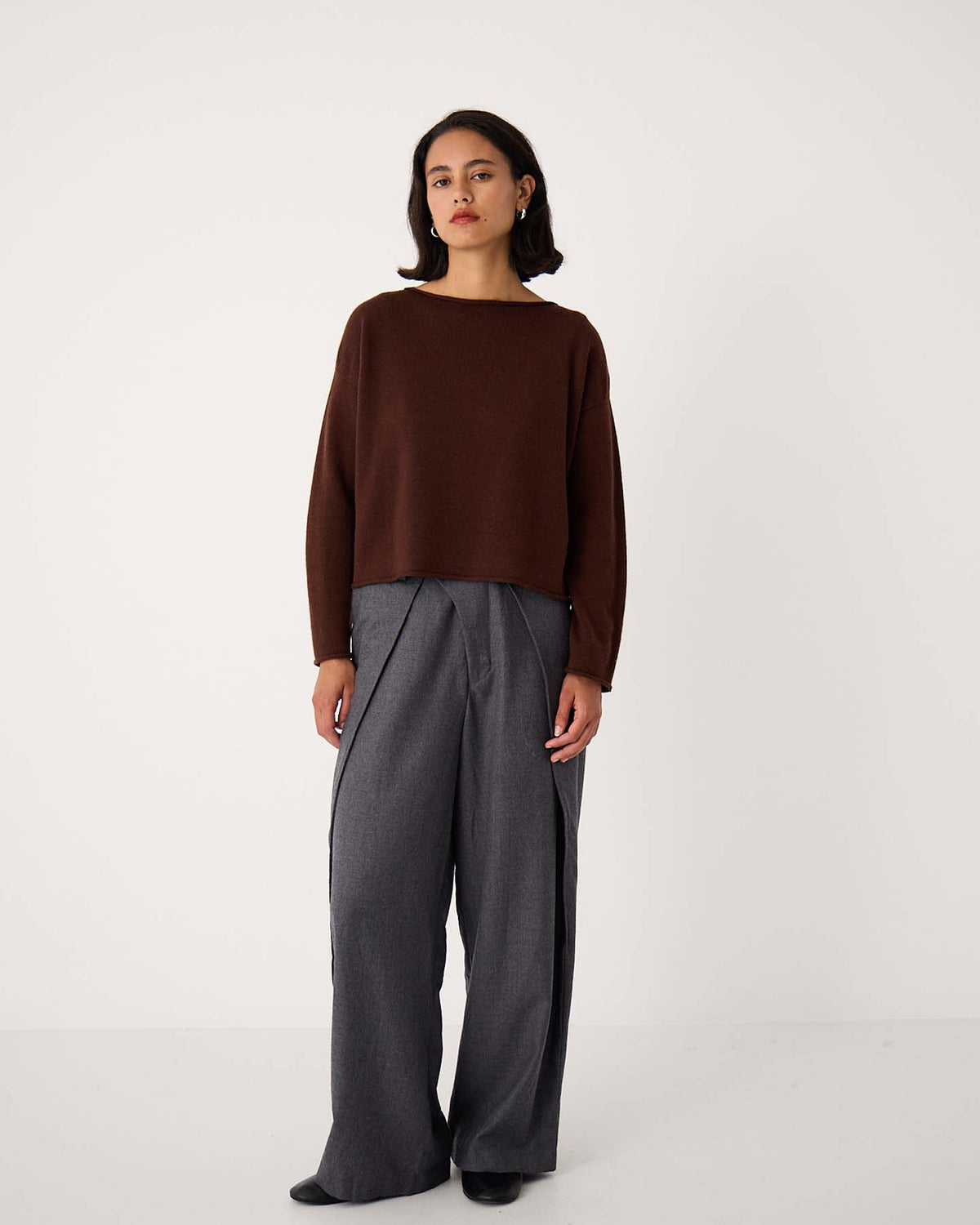 A woman stands against a plain white background wearing the Doll's Jumper – Chestnut, a relaxed fit cropped brown sweater, paired with wide-leg gray trousers and black shoes—an effortless look ideal for petite clothing lovers.