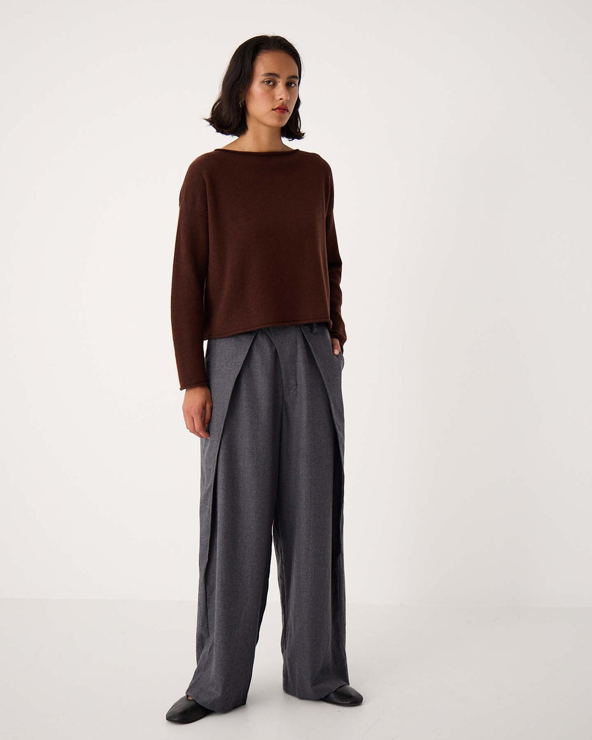 A woman stands against a plain white background wearing the Doll's Jumper – Chestnut, paired with relaxed fit, wide-leg gray pleated trousers and black shoes.