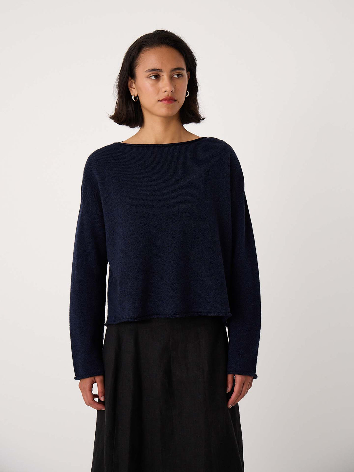 A woman stands against a plain background, wearing the Doll's Jumper – Ink, a navy blue long-sleeve relaxed fit sweater, paired with a black skirt and looking slightly to the side.