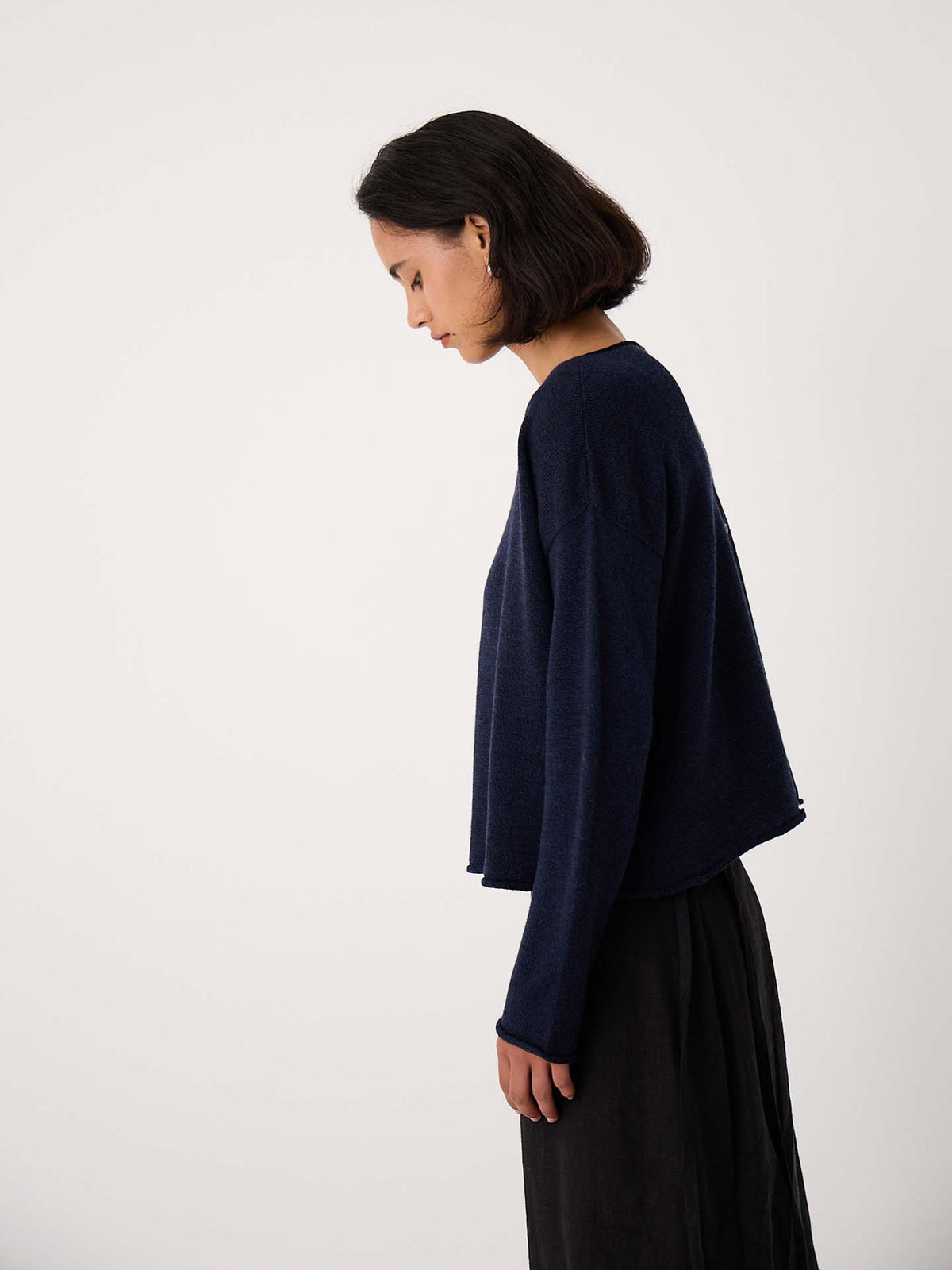 A woman with short dark hair stands in profile against a plain light background, wearing the Doll's Jumper – Ink, a navy long-sleeve top in a relaxed fit, paired with a black skirt.