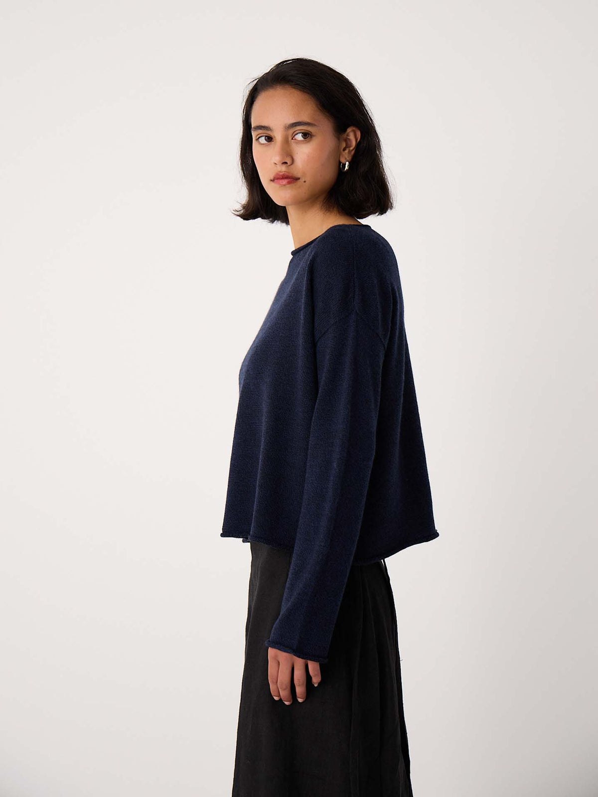 A woman with short dark hair wears the Doll's Jumper – Ink, a relaxed-fit navy long-sleeve top, paired with a black skirt as she stands sideways against a plain white background.