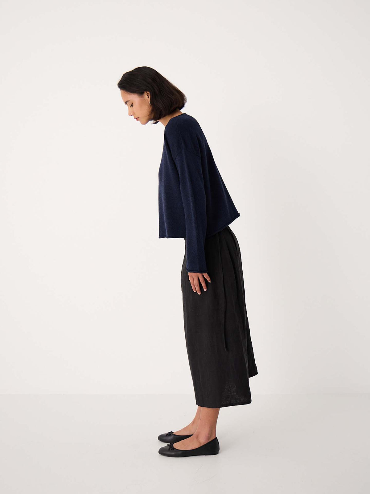 A woman stands in profile against a plain background, wearing the Doll's Jumper – Ink, black wide-leg pants, and black flats, with her head slightly bowed.