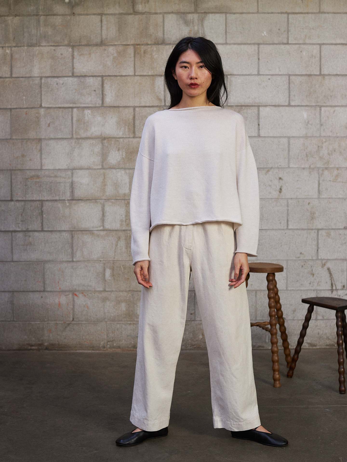 A petite woman stands indoors by a gray cinder block wall, wearing the Doll's Jumper – Ivory and matching wide-leg pants with black flats. Two wooden stools are placed behind her.