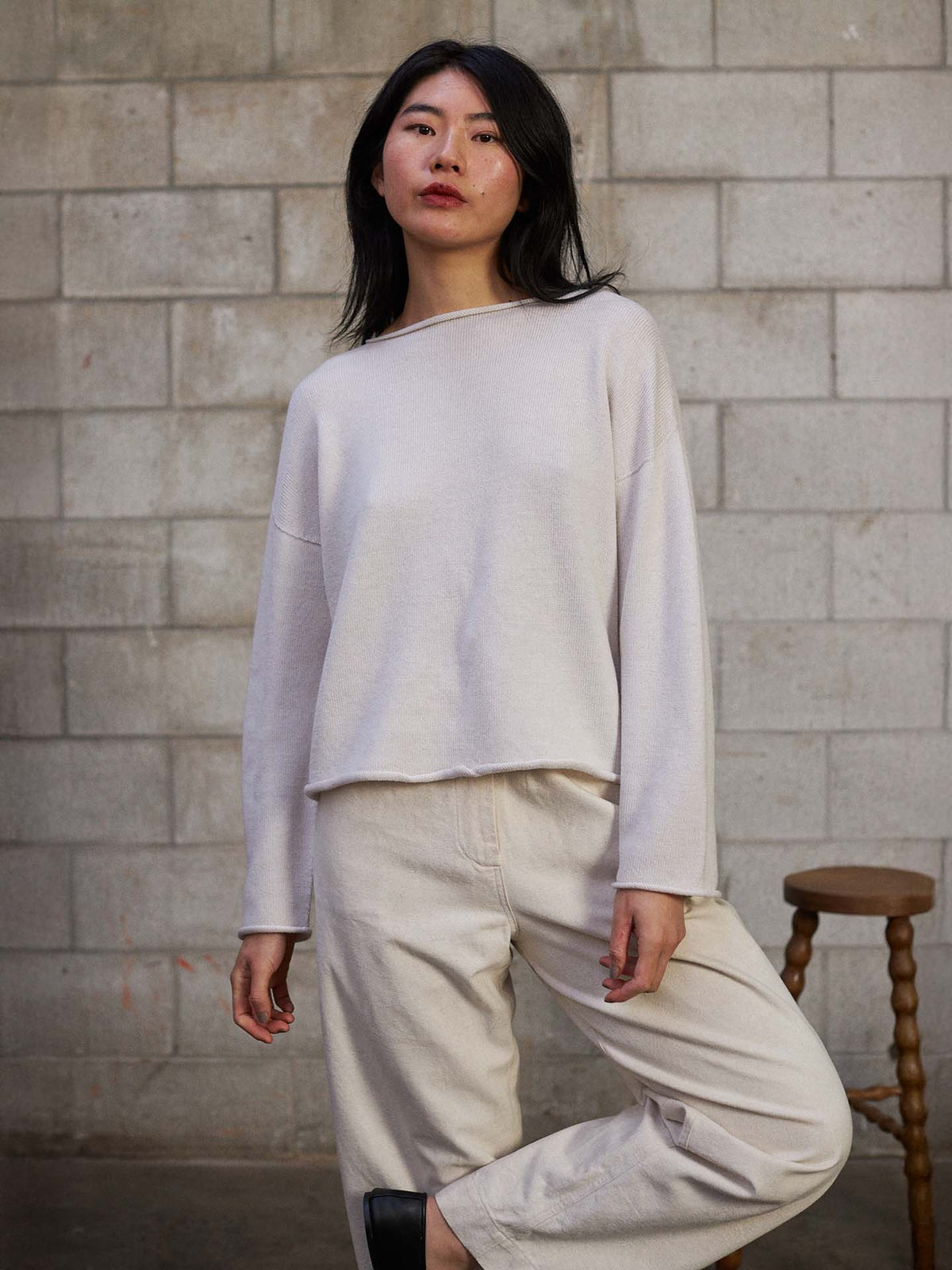 A petite person wears the Doll's Jumper – Ivory, a relaxed fit long-sleeve top and pants set, standing indoors by a gray cinder block wall with a wooden stool in the background.