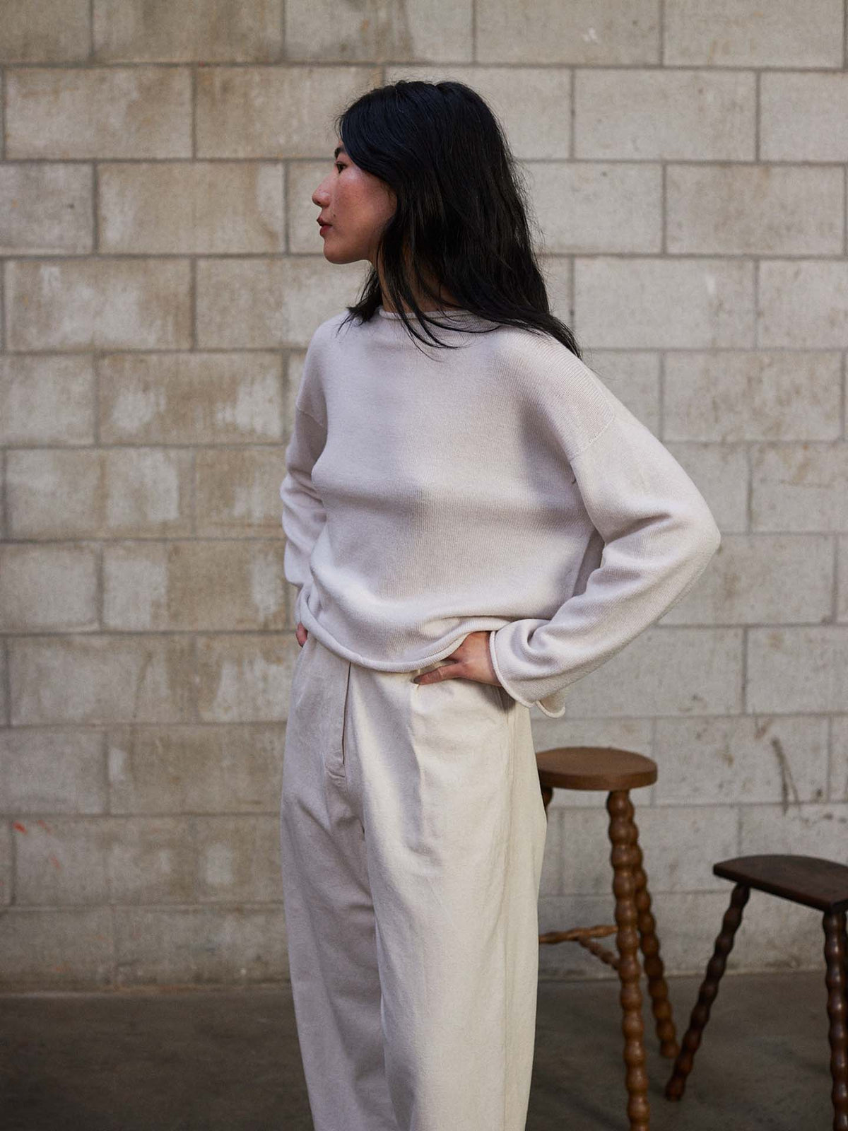 A petite woman stands sideways with hands on hips wearing the Doll's Jumper – Ivory, a relaxed fit sweater and pants set, in front of a concrete block wall next to two wooden stools.