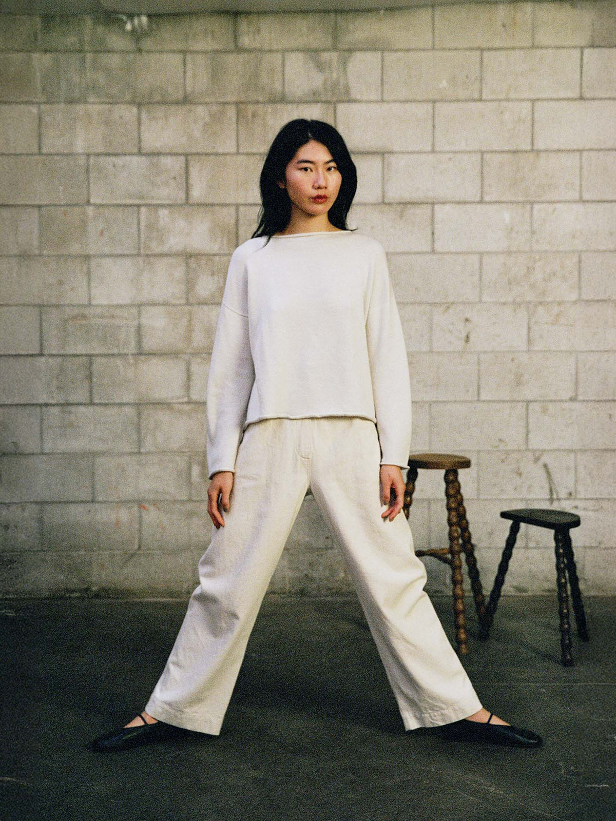 Wearing the Doll's Jumper – Ivory, a person in relaxed-fit, light clothing stands with legs apart before a concrete block wall, with two wooden stools visible in the background.