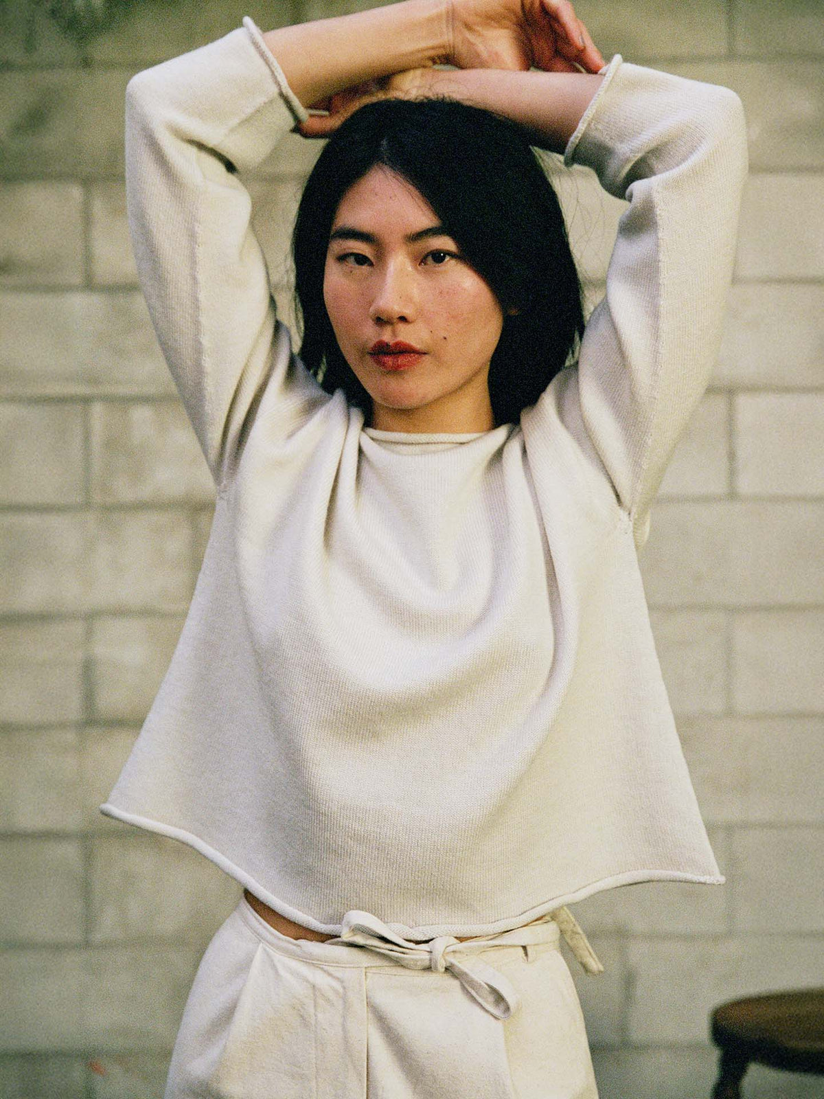 A petite person wears the Doll's Jumper – Ivory, a relaxed-fit, light-colored long-sleeve top and pants, standing against a cinder block wall with arms raised overhead.