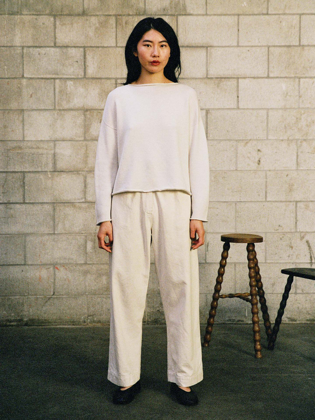 A woman stands facing forward against a concrete block wall, wearing the Doll's Jumper – Ivory, a relaxed fit, light-colored long-sleeve top with wide-leg pants. Two stools are visible in the background.