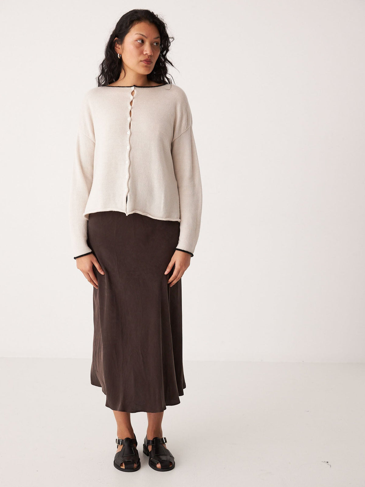 A woman wears the Francie Doll&#39;s Reversible Cardigan in Ivory &amp; Black against a white background, paired with a dark brown midi skirt and black sandals. Check our size guide to find your perfect fit in petite styles.