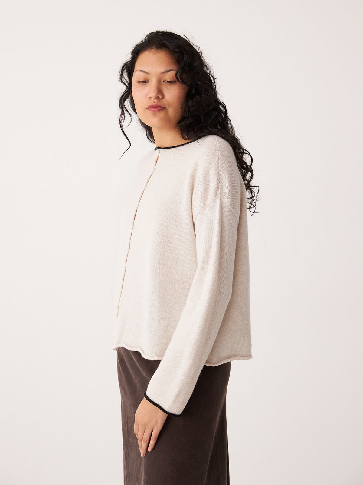 A woman with long curly hair wears the Francie Doll&#39;s Reversible Cardigan in ivory and black, styled cropped with black trim, paired with a dark skirt, standing against a plain white background.