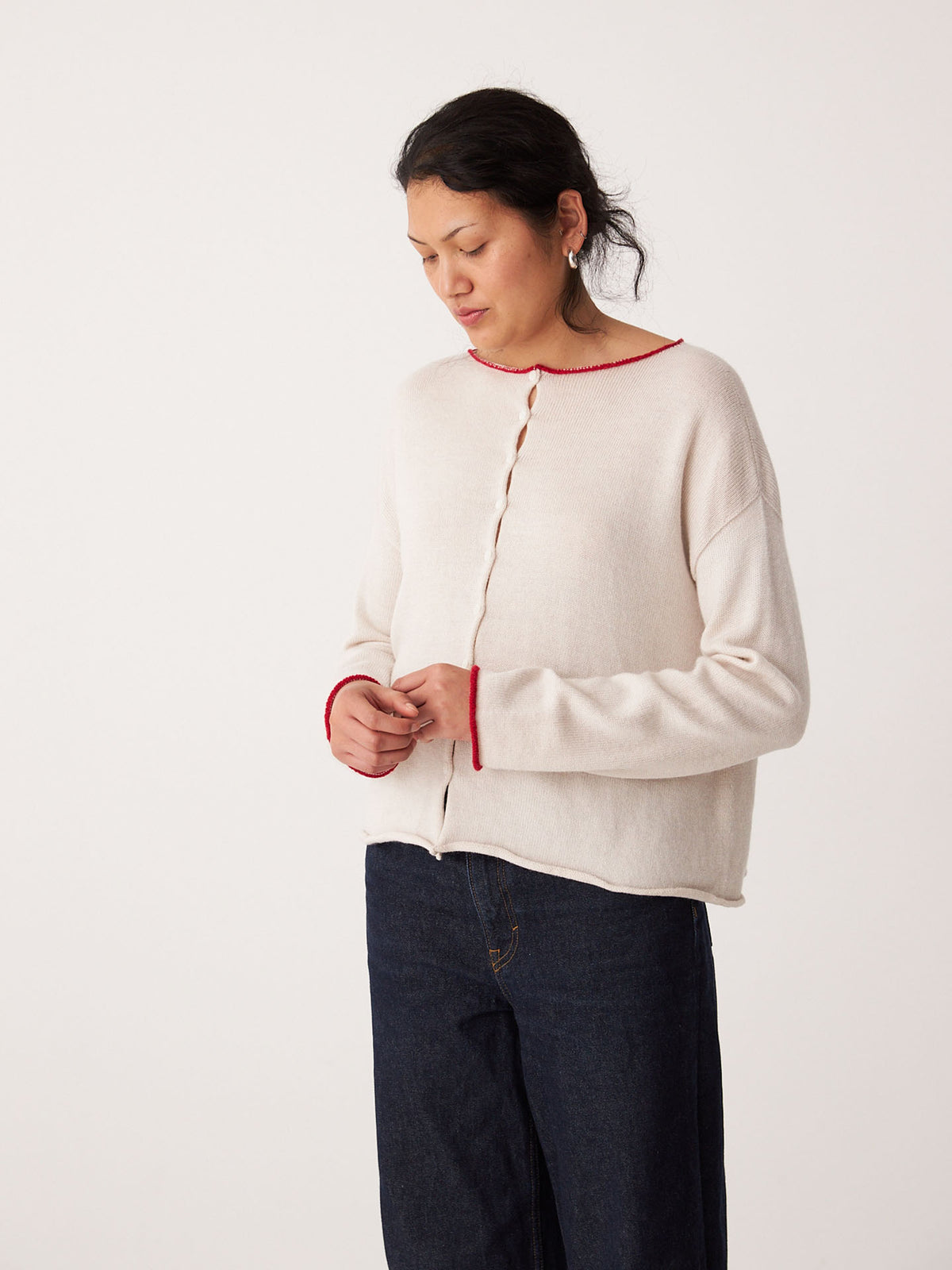 A person models the Francie Doll&#39;s Reversible Cardigan in ivory and red, paired with dark jeans, standing against a plain white background and looking down.