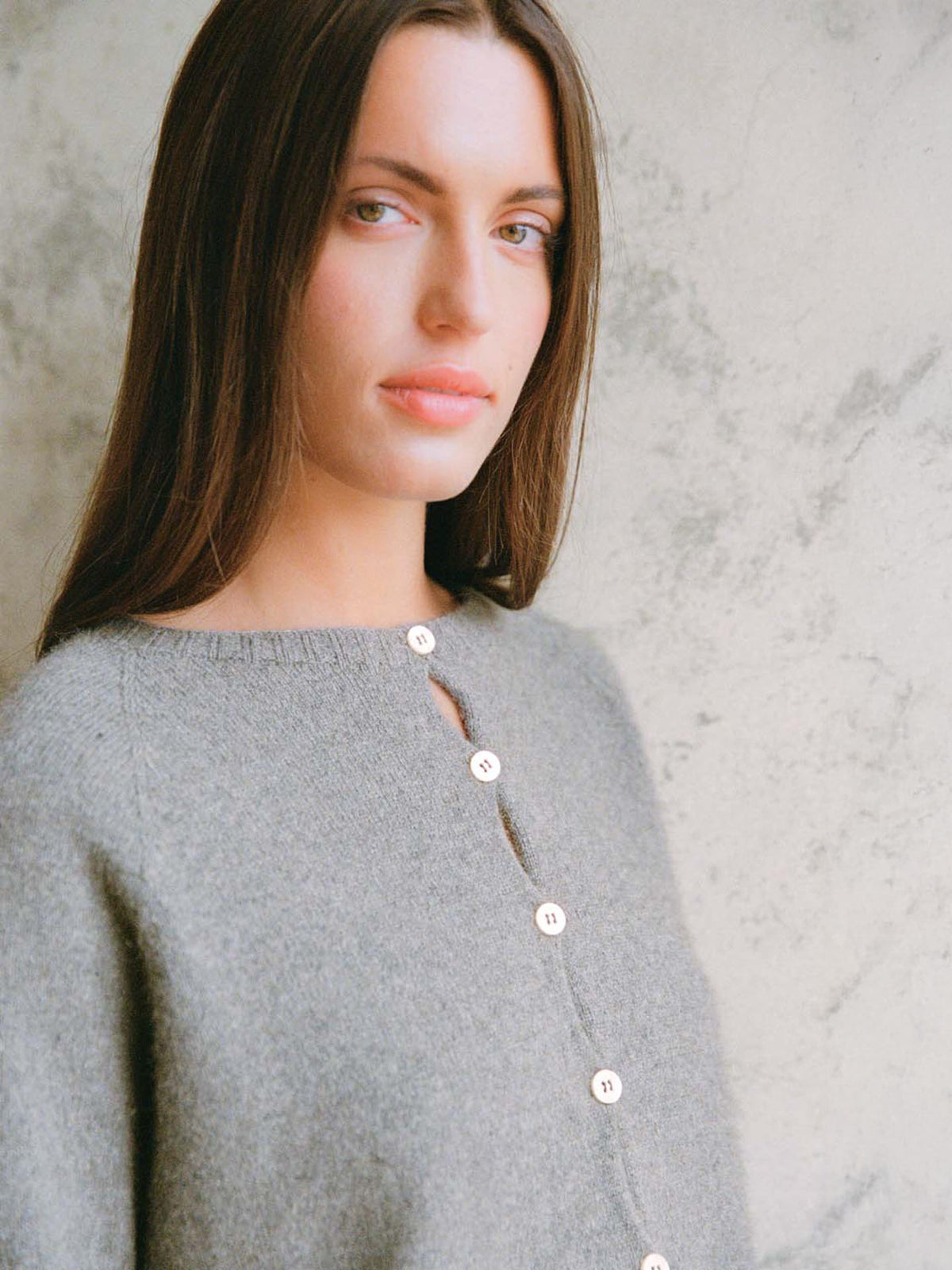 A petite woman with long brown hair wears the Francie Dusk Cardigan in Granite, a relaxed-fit buttoned sweater, standing before a light-textured wall and looking at the camera.