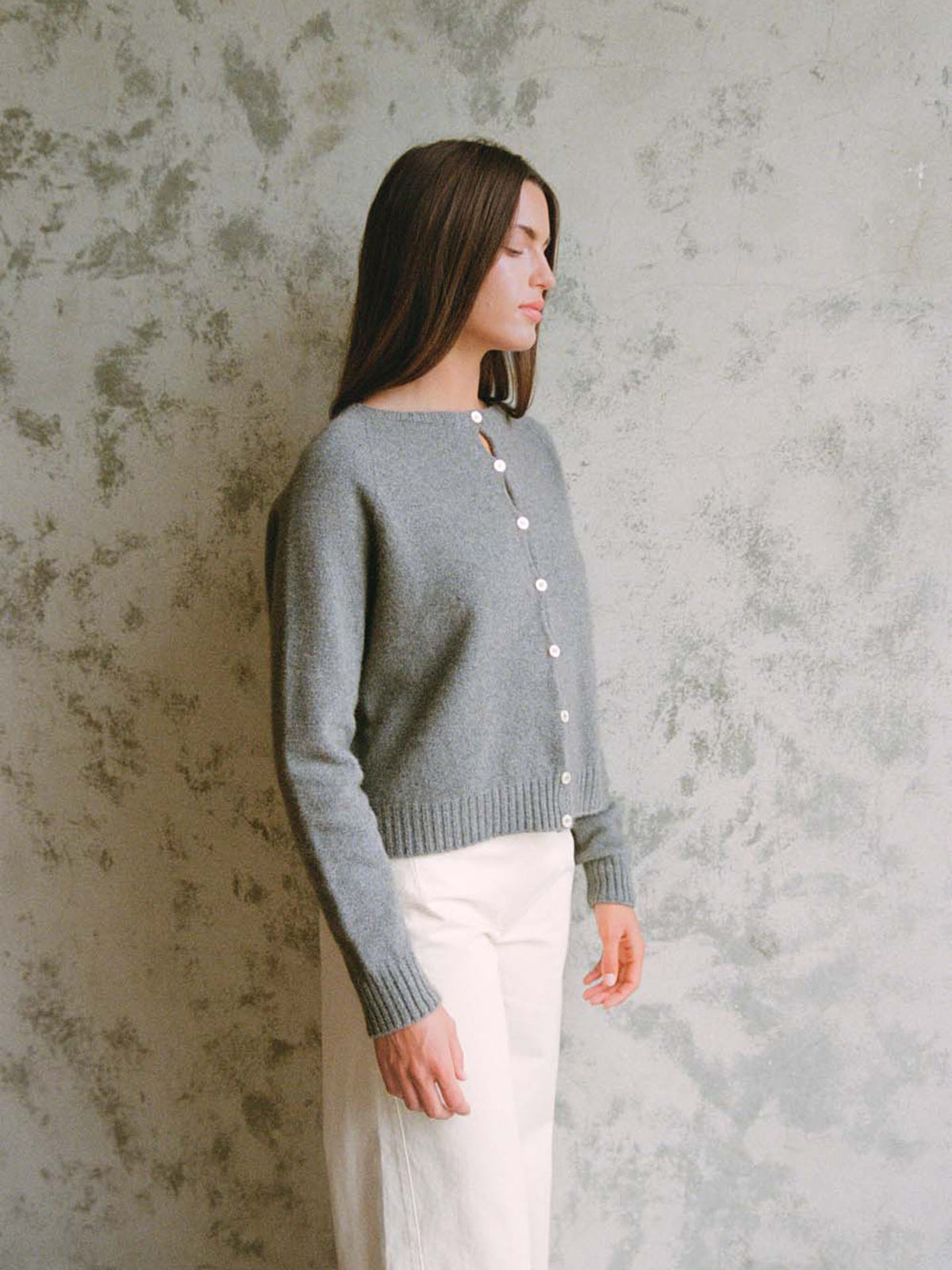 A petite woman with long brown hair, wearing Francie&#39;s Dusk Cardigan in Granite and white pants, stands sideways against a textured light gray wall.