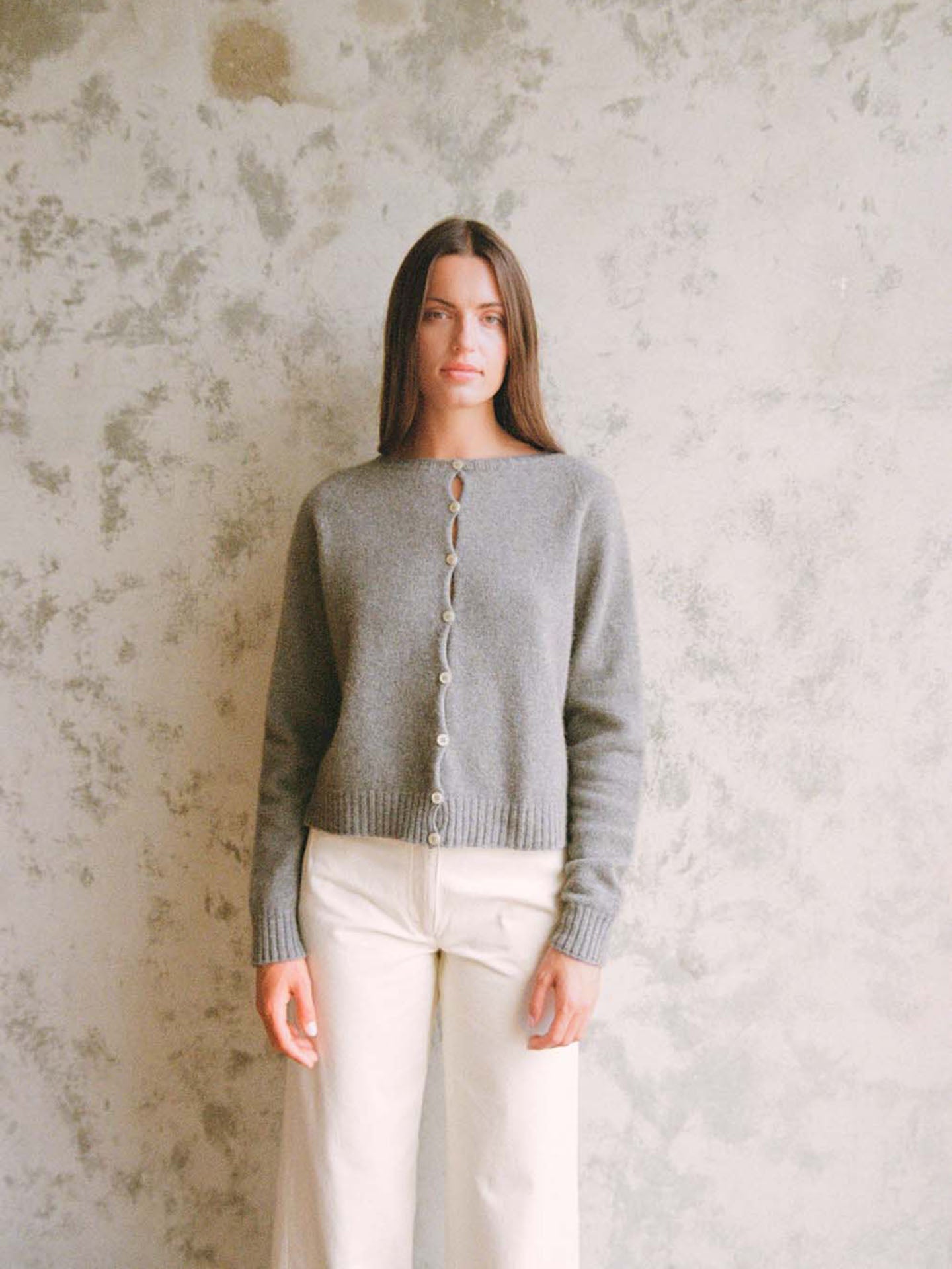 A woman stands against a textured light grey wall, wearing Francie's Dusk Cardigan – Granite in a relaxed fit, paired with cream-colored pants.