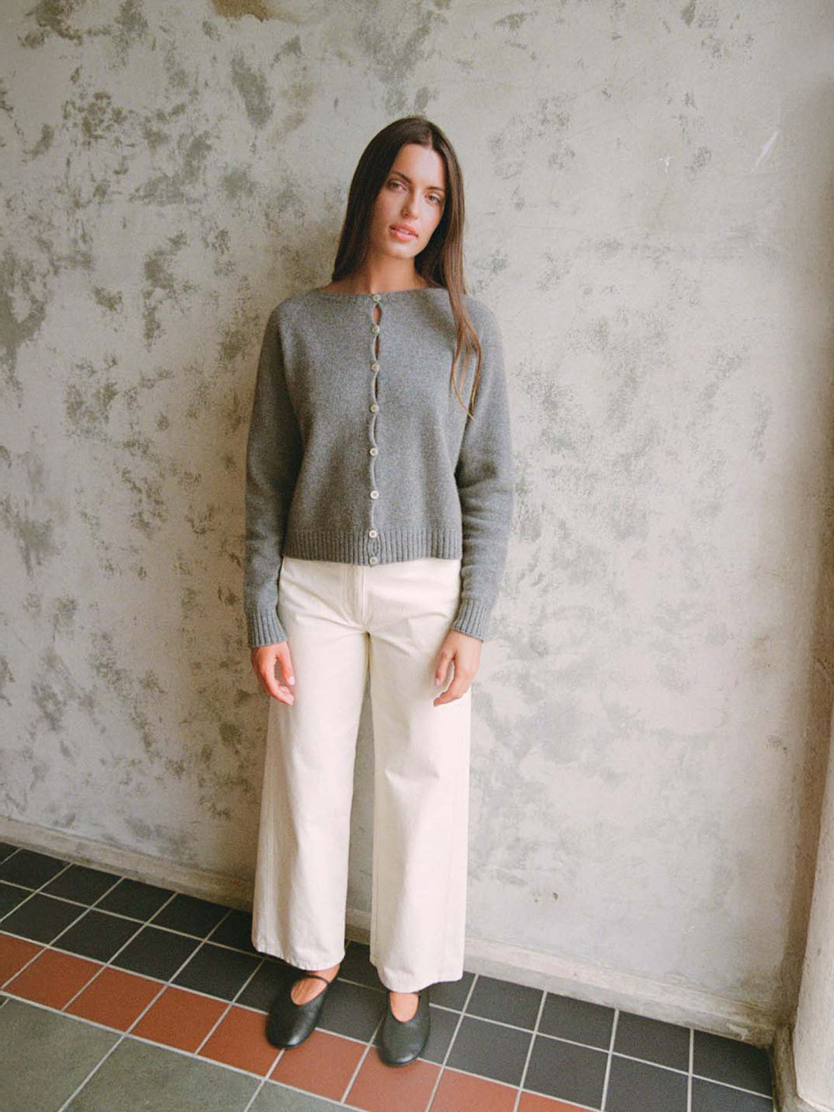 A woman stands indoors against a textured wall, wearing Francie&#39;s Dusk Cardigan in Granite, styled with wide-leg white pants and black shoes.