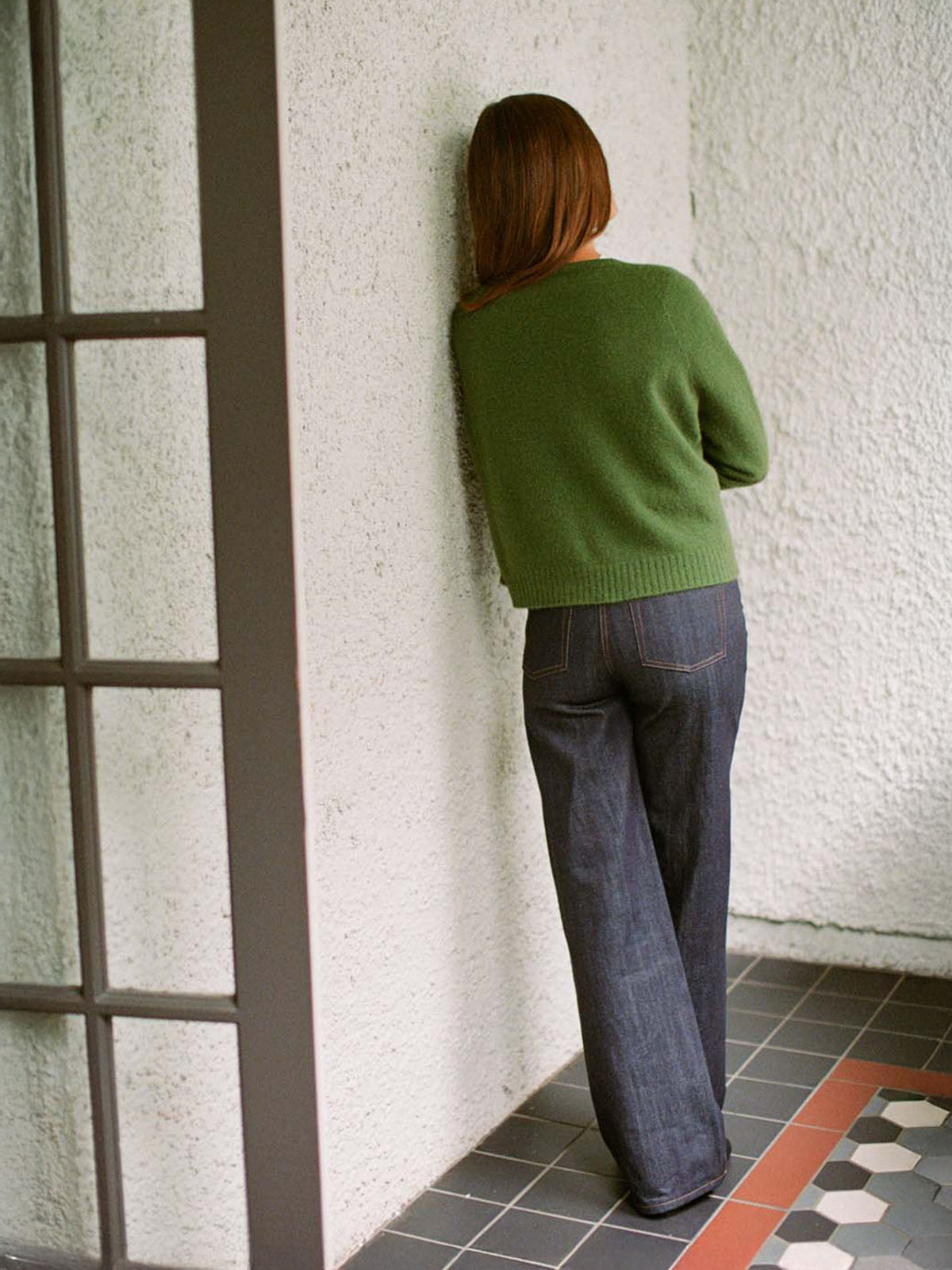 A petite person with long hair wears the Francie Dusk Cardigan in Herb Green, styled with jeans, standing before a textured white wall in a tiled indoor setting.