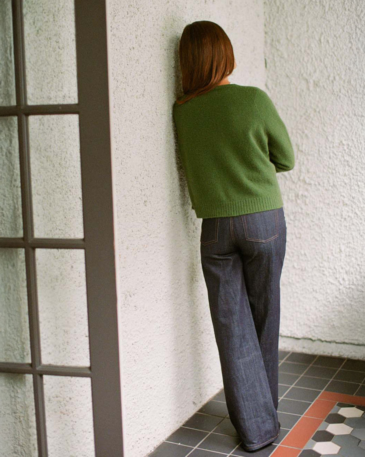 A petite person with long hair wears the Francie Dusk Cardigan in Herb Green, styled with jeans, standing before a textured white wall in a tiled indoor setting.