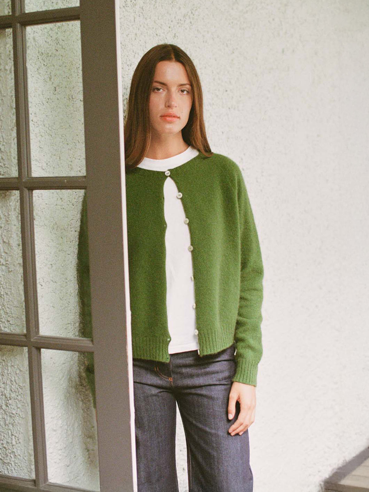 A person with straight brown hair stands indoors by a window, wearing Francie&#39;s Dusk Cardigan in Herb Green over a white shirt and blue jeans, looking directly at the camera.
