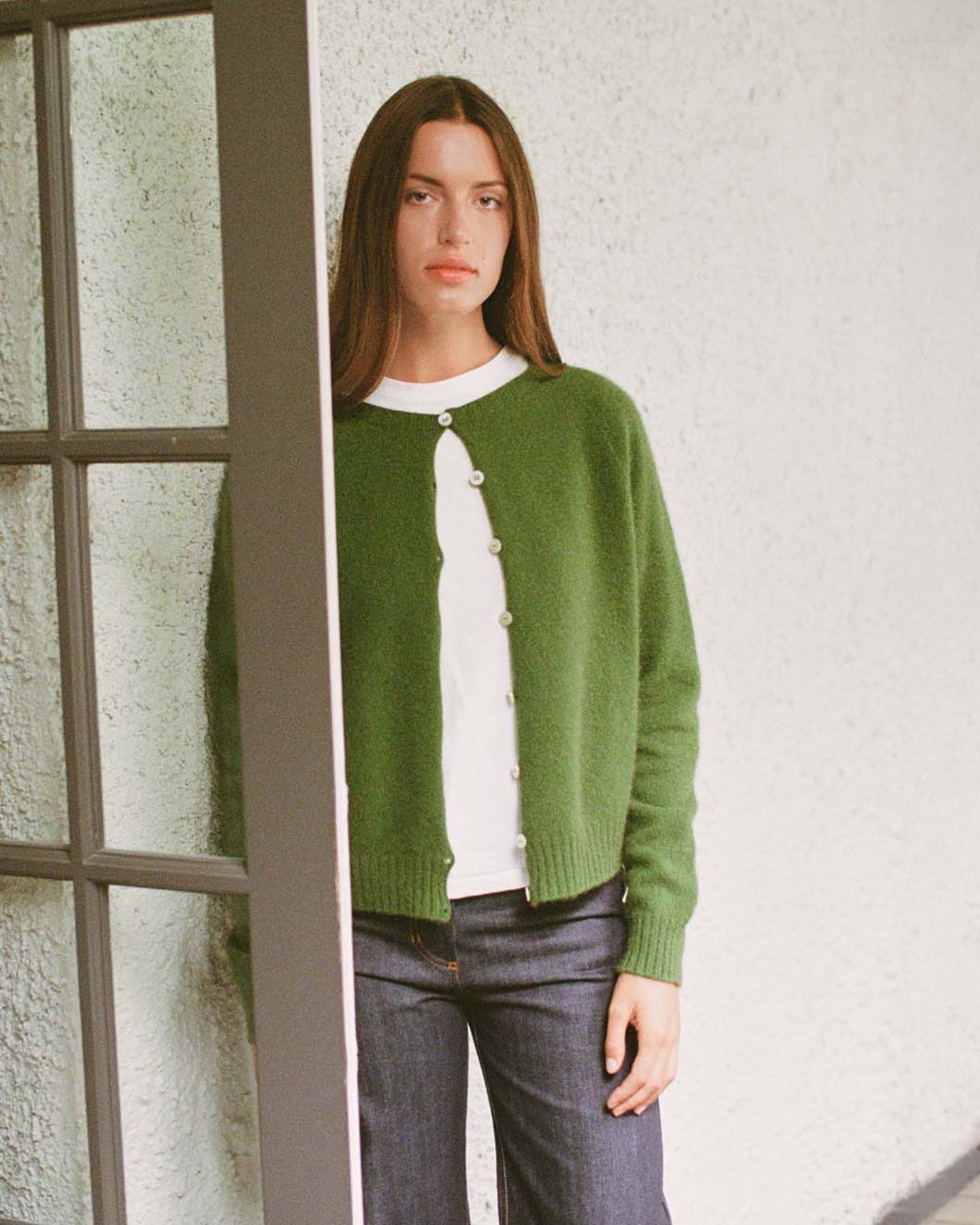 A person with straight brown hair stands indoors by a window, wearing Francie's Dusk Cardigan in Herb Green over a white shirt and blue jeans, looking directly at the camera.