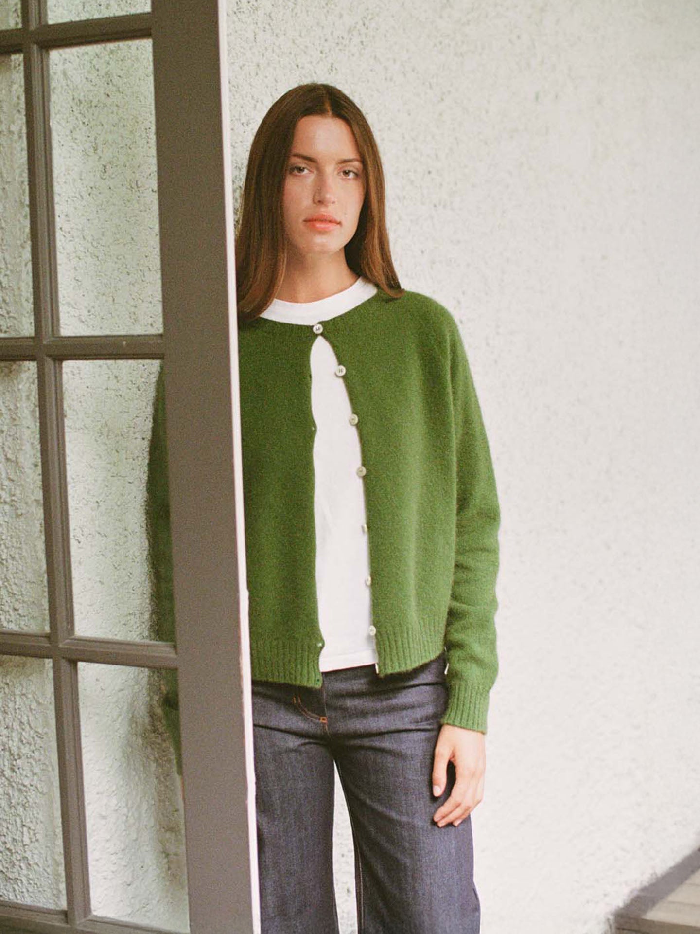 A person with straight brown hair stands indoors by a window, wearing Francie's Dusk Cardigan in Herb Green over a white shirt and blue jeans, looking directly at the camera.
