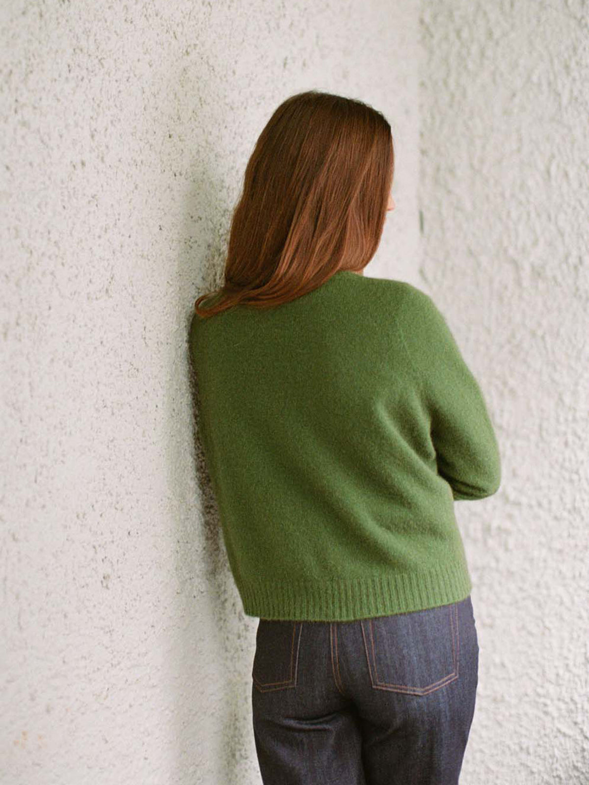 A petite person with long brown hair wears Francie&#39;s Dusk Cardigan in Herb Green, paired with dark jeans, standing back to the camera against a textured white wall.