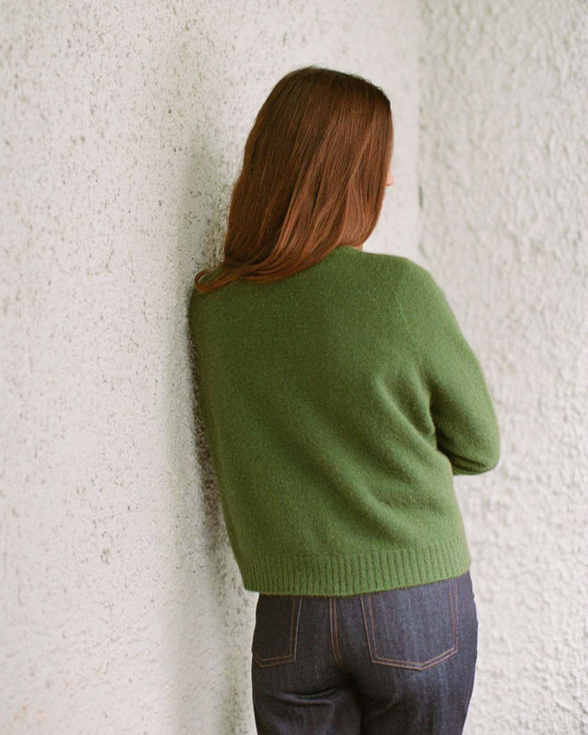 A petite person with long brown hair wears Francie's Dusk Cardigan in Herb Green, paired with dark jeans, standing back to the camera against a textured white wall.