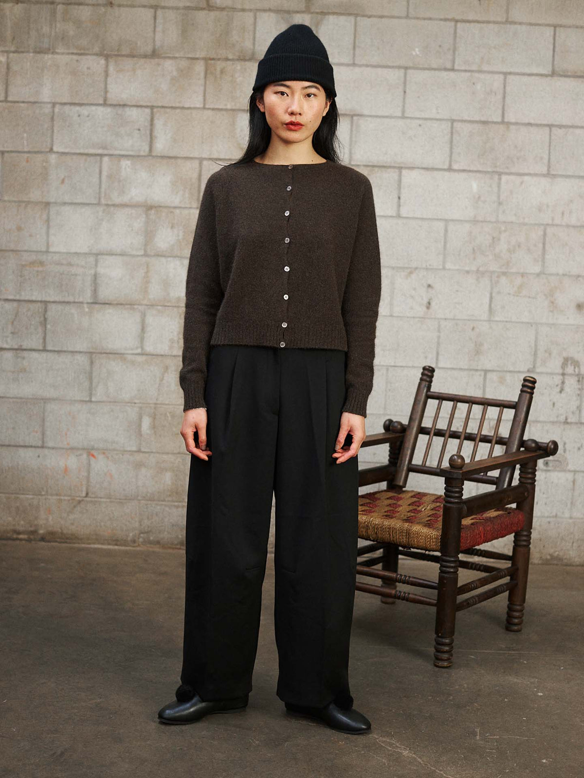 A woman wearing a black beanie, the Dusk Cardigan – Truffle in a relaxed fit, and black wide-leg pants stands indoors before a stone wall and wooden chair.