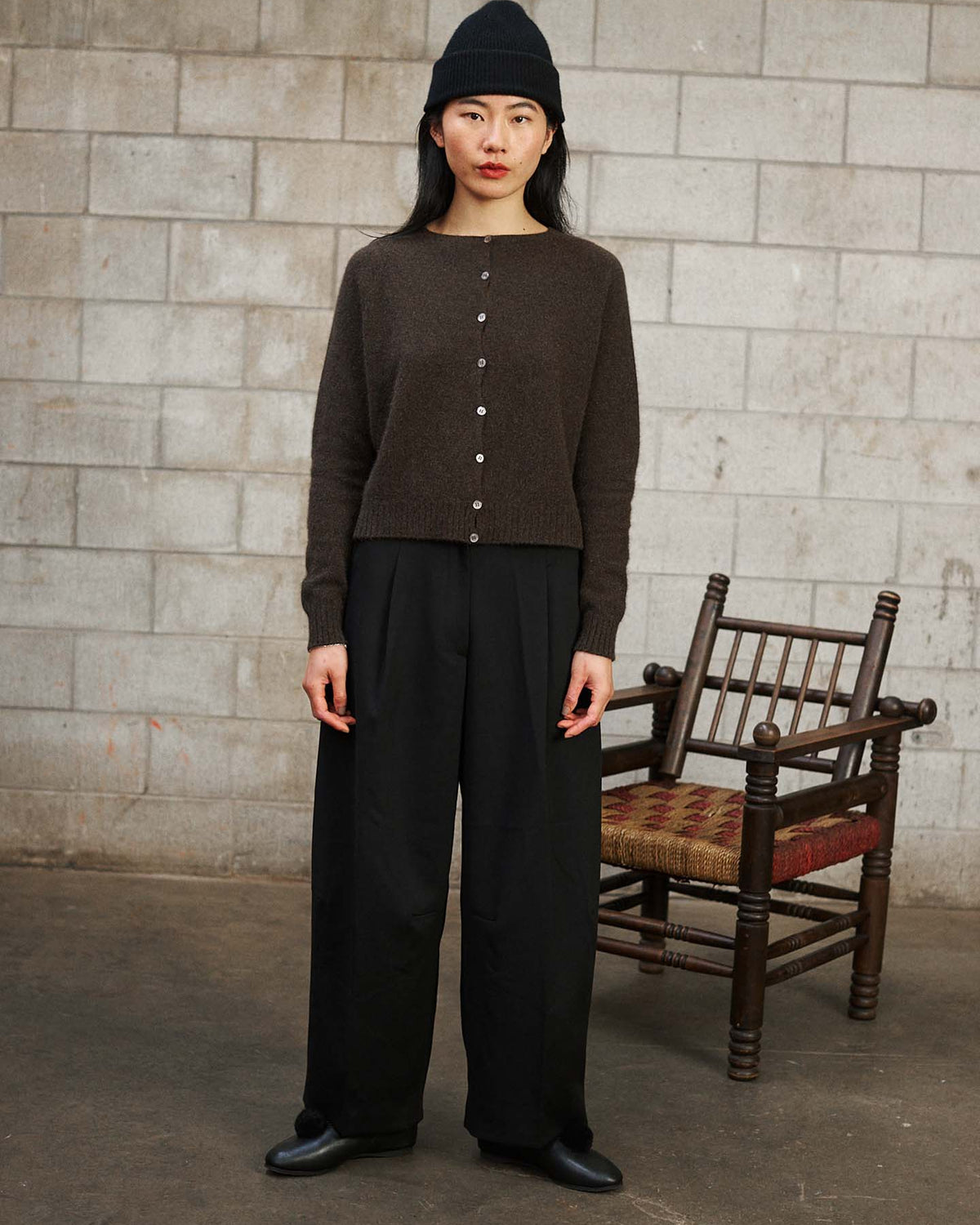 A woman wearing a black beanie, the Dusk Cardigan – Truffle in a relaxed fit, and black wide-leg pants stands indoors before a stone wall and wooden chair.