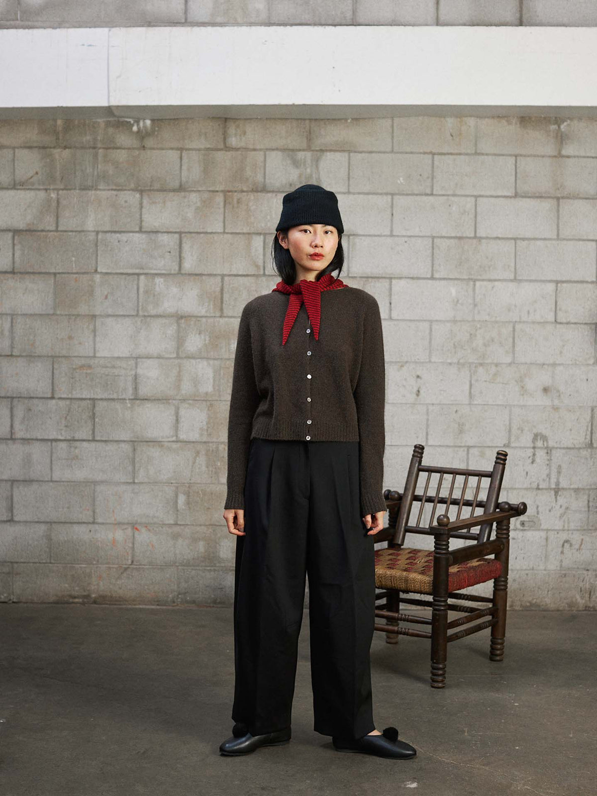 A woman wears the Dusk Cardigan in Truffle, paired with black pants, a black hat, and a red scarf, standing indoors on a concrete floor before a cinder block wall with a wooden chair behind her.