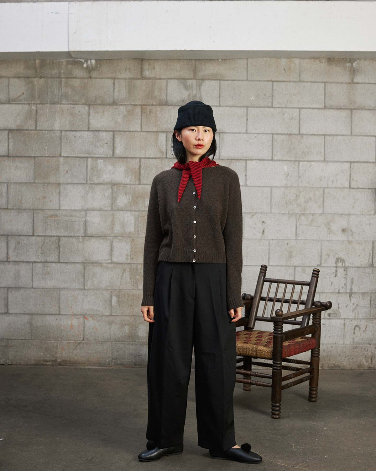 A woman wears the Dusk Cardigan in Truffle, paired with black pants, a black hat, and a red scarf, standing indoors on a concrete floor before a cinder block wall with a wooden chair behind her.