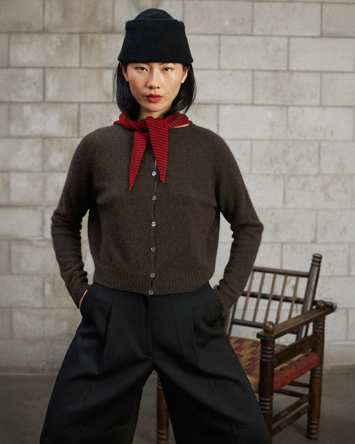A woman stands indoors wearing a black beanie, red scarf, Dusk Cardigan – Truffle in a relaxed fit, and black pants, with her hands in her pockets and an empty wooden chair behind her.
