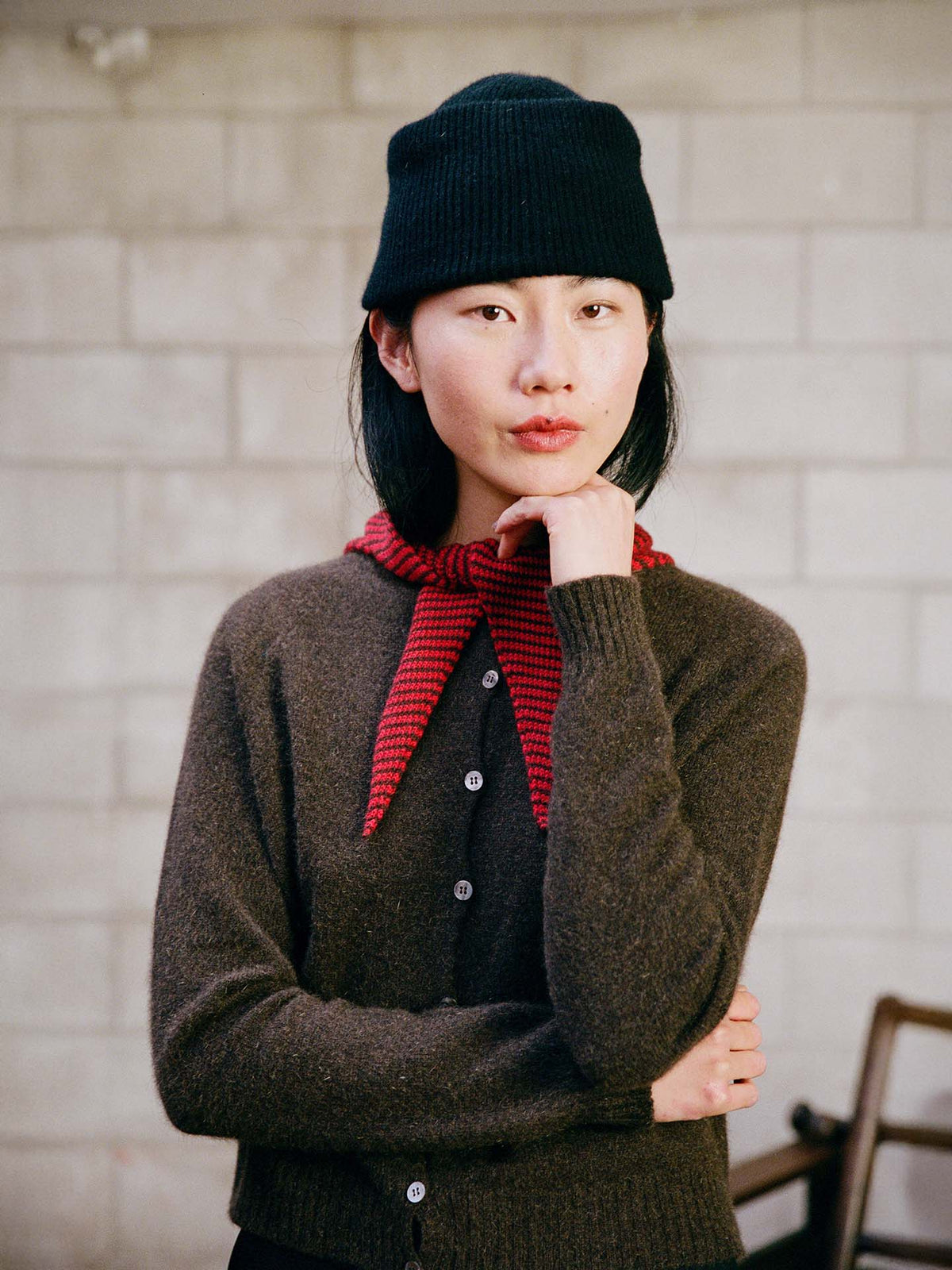 A person in a relaxed fit Dusk Cardigan – Truffle, black knit hat, and red scarf stands indoors against a light brick wall, resting their chin on one hand.