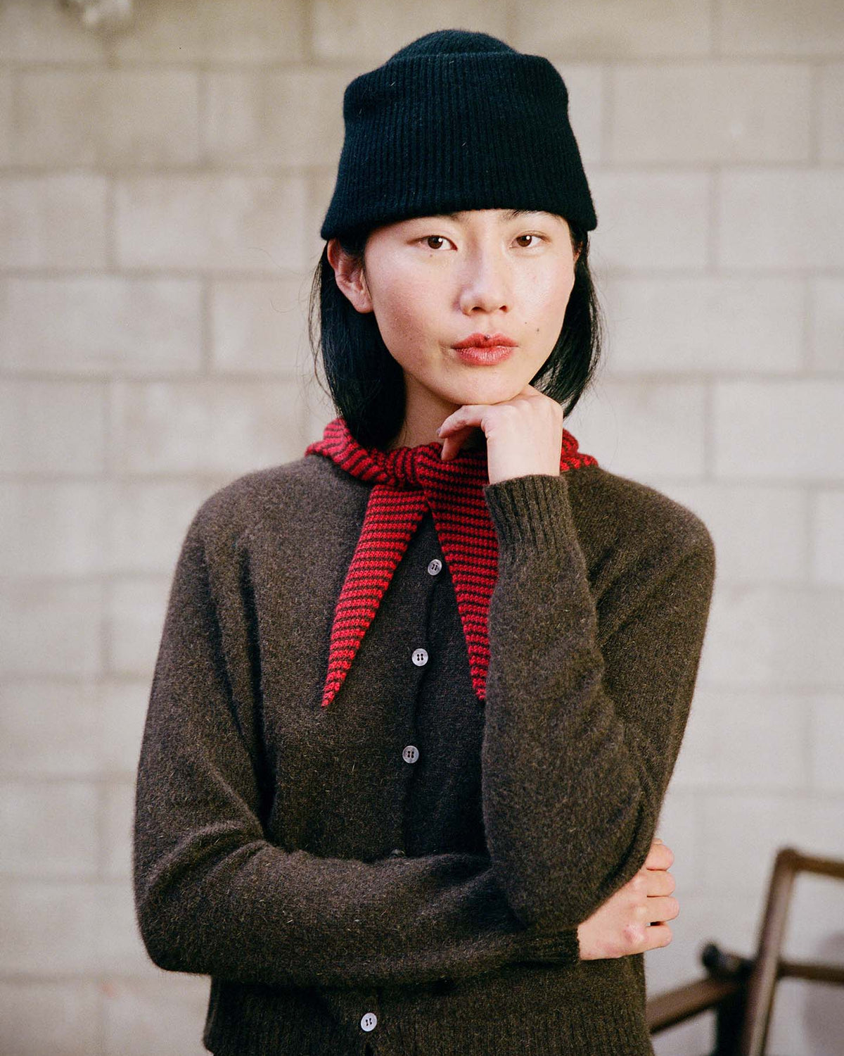 A person in a relaxed fit Dusk Cardigan – Truffle, black knit hat, and red scarf stands indoors against a light brick wall, resting their chin on one hand.