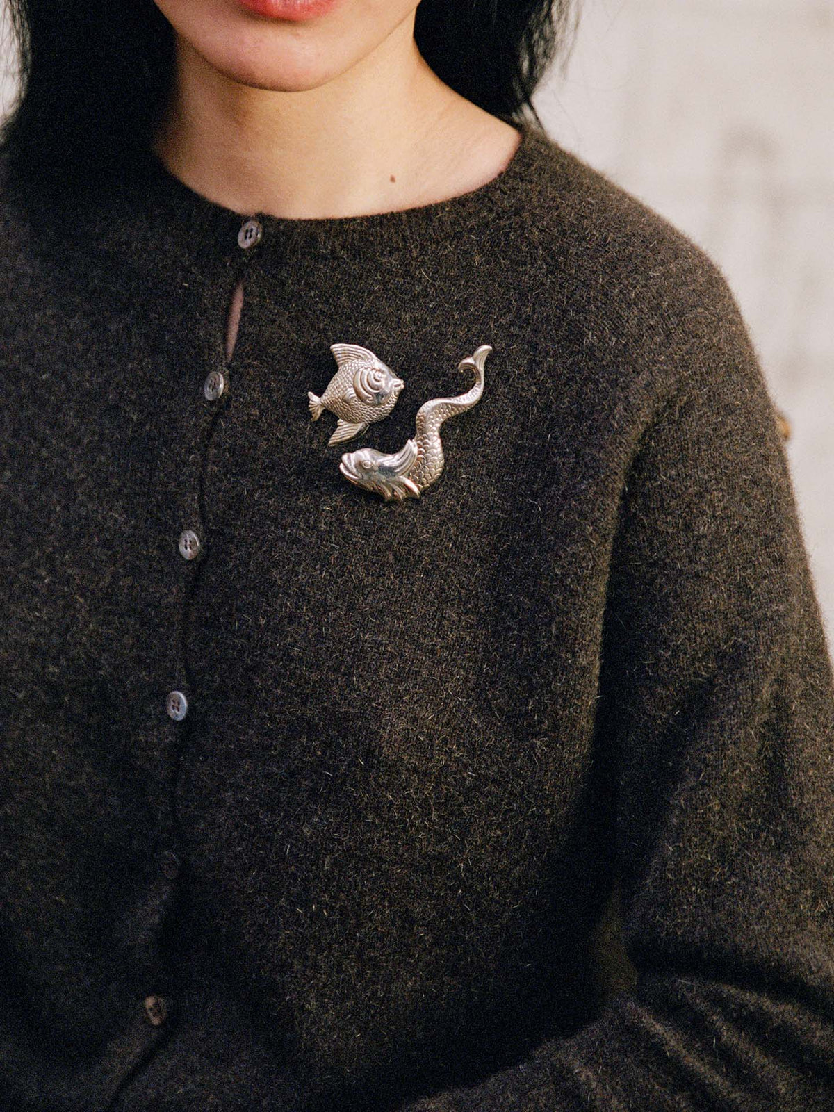 A person wearing the Dusk Cardigan – Truffle, a relaxed-fit dark brown buttoned sweater, styled with two silver brooches shaped like a fish and a snake on the chest.