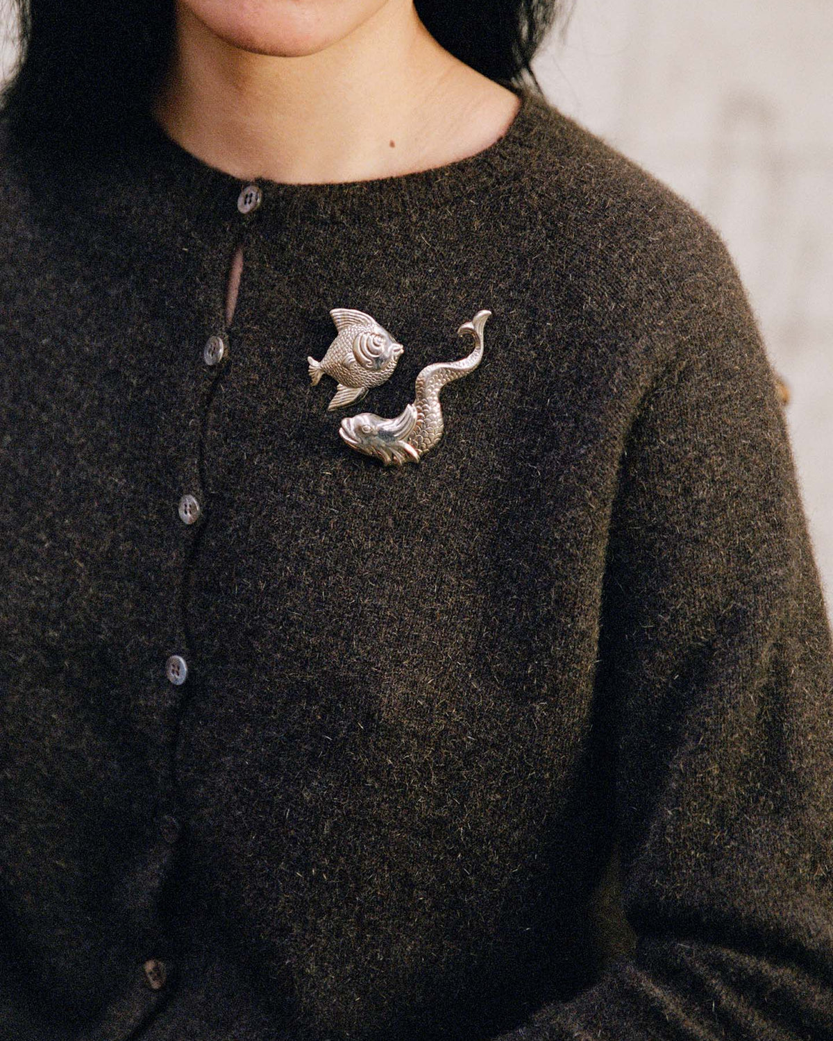 A person wearing the Dusk Cardigan – Truffle, a relaxed-fit dark brown buttoned sweater, styled with two silver brooches shaped like a fish and a snake on the chest.