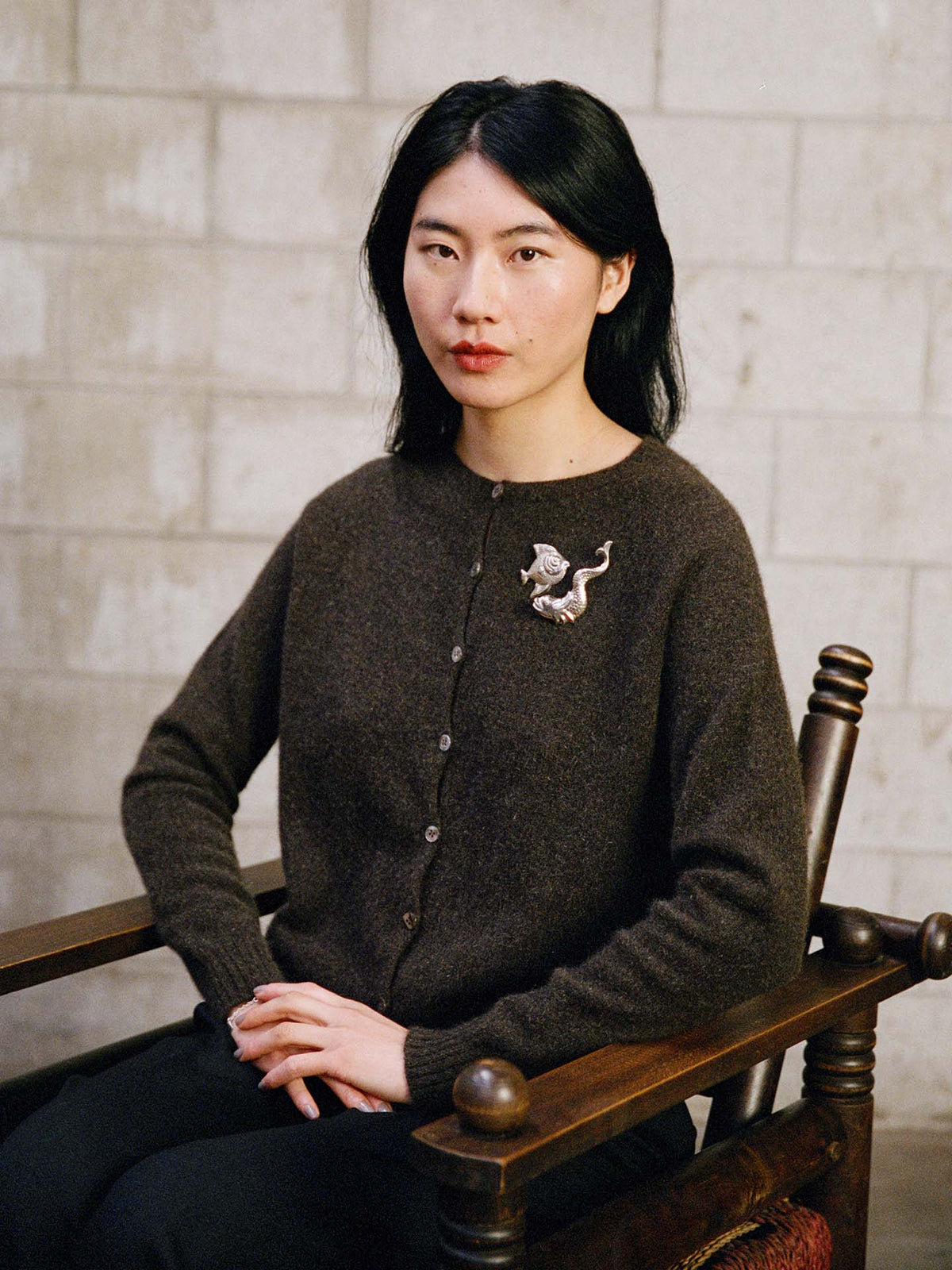 A woman with long black hair sits on a wooden chair against a light brick wall, wearing the Dusk Cardigan – Truffle in a relaxed fit and accessorized with a silver creature-shaped brooch.