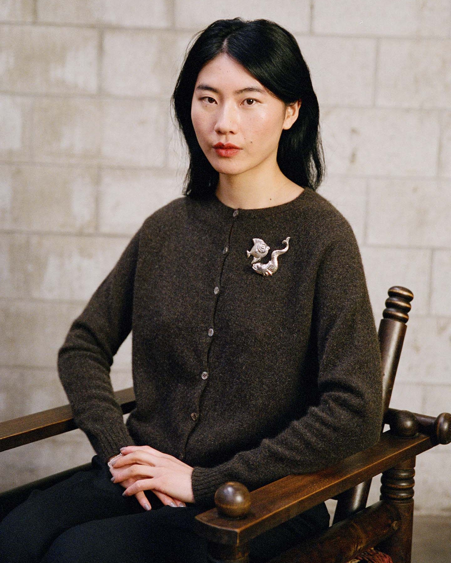 A woman with long black hair sits on a wooden chair against a light brick wall, wearing the Dusk Cardigan – Truffle in a relaxed fit and accessorized with a silver creature-shaped brooch.