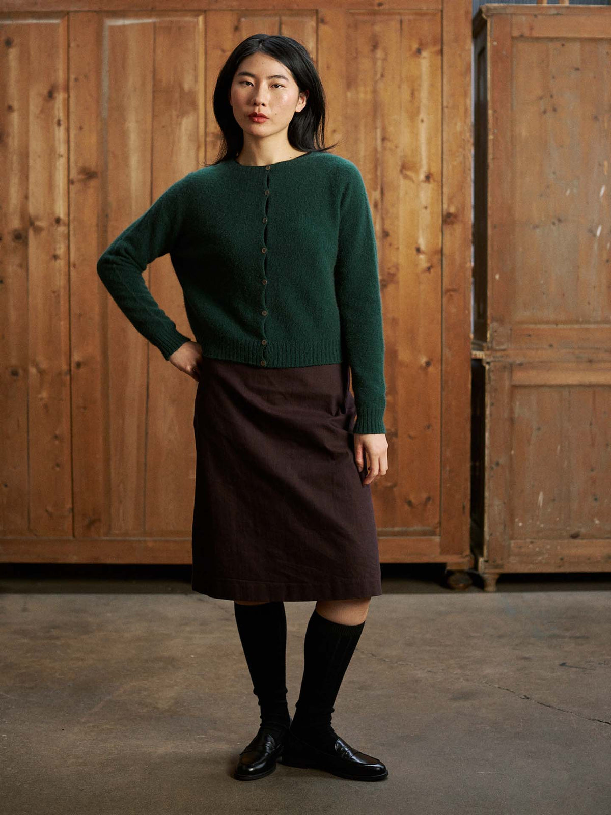 A woman stands indoors against a wooden background, wearing the Dusk Cardigan in Verdant Green, paired with a brown skirt, black knee-high socks, and black shoes.