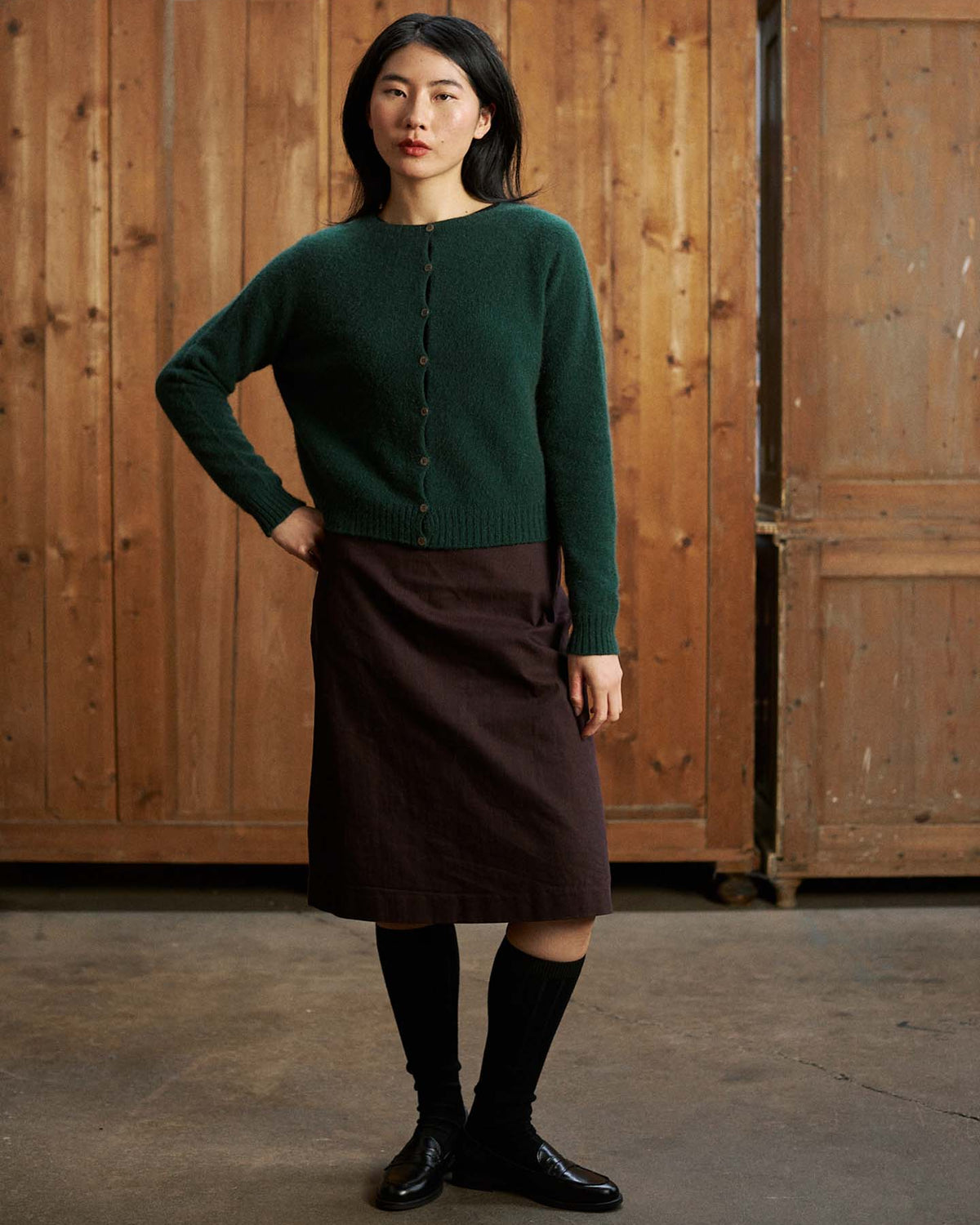 A woman stands indoors against a wooden background, wearing the Dusk Cardigan in Verdant Green, paired with a brown skirt, black knee-high socks, and black shoes.