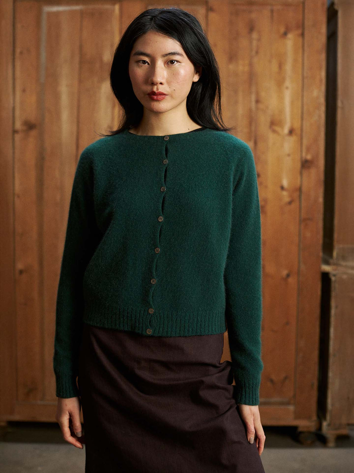 A woman with long dark hair stands indoors by a wooden wall, wearing the Dusk Cardigan in Verdant Green—a relaxed-fit, buttoned sweater—paired with a dark skirt.