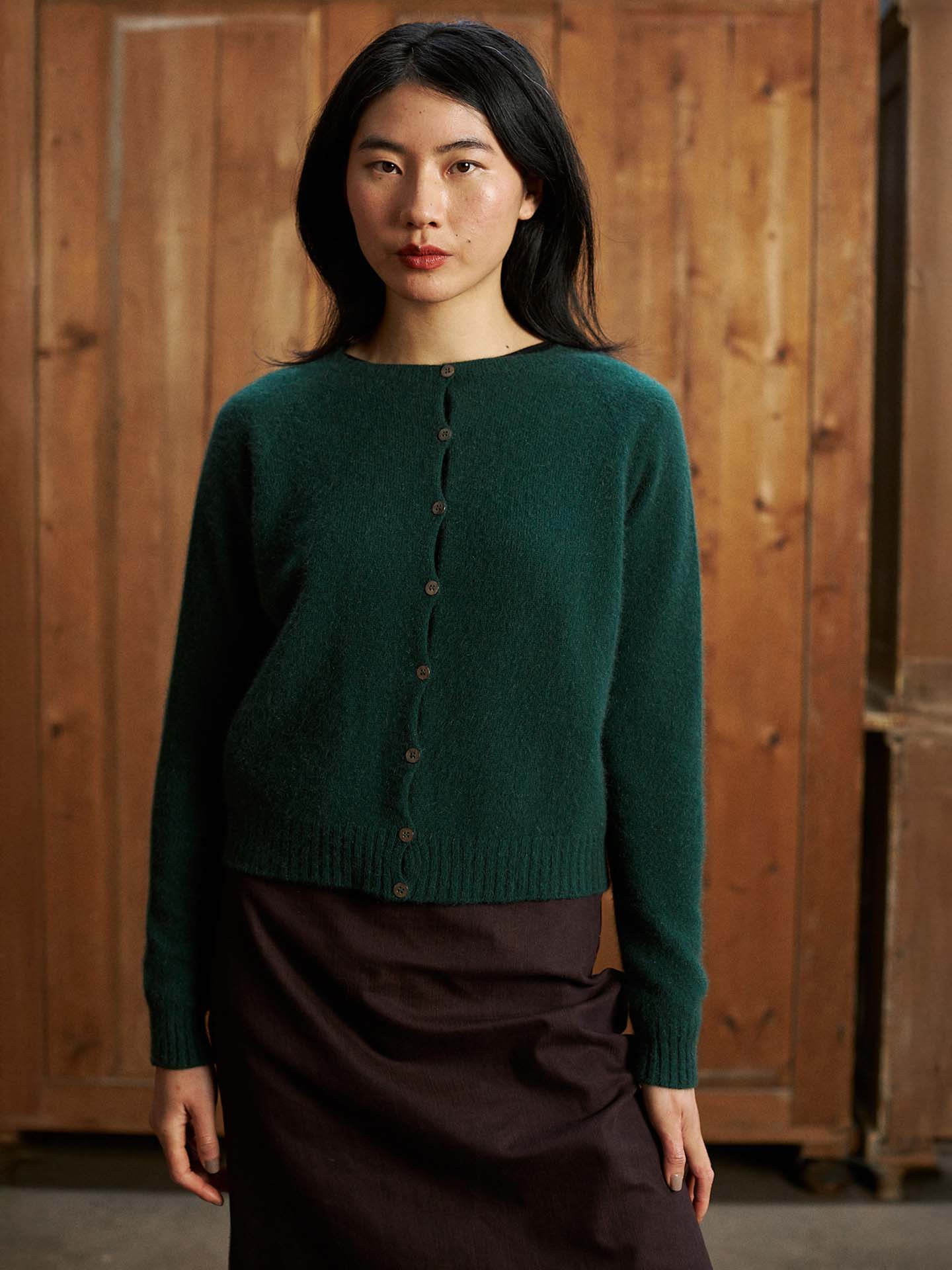 A woman with long dark hair stands indoors by a wooden wall, wearing the Dusk Cardigan in Verdant Green—a relaxed-fit, buttoned sweater—paired with a dark skirt.