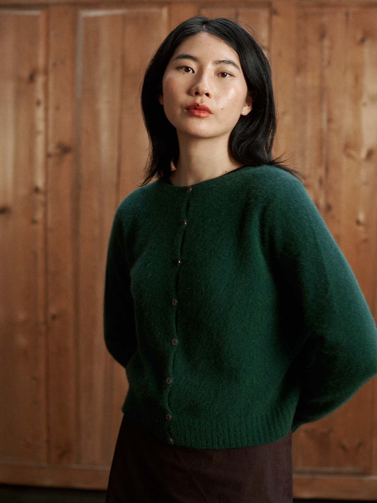 A woman with medium-length dark hair wears the Dusk Cardigan in Verdant Green, a relaxed fit buttoned sweater, as she stands before a wooden wall and looks at the camera.